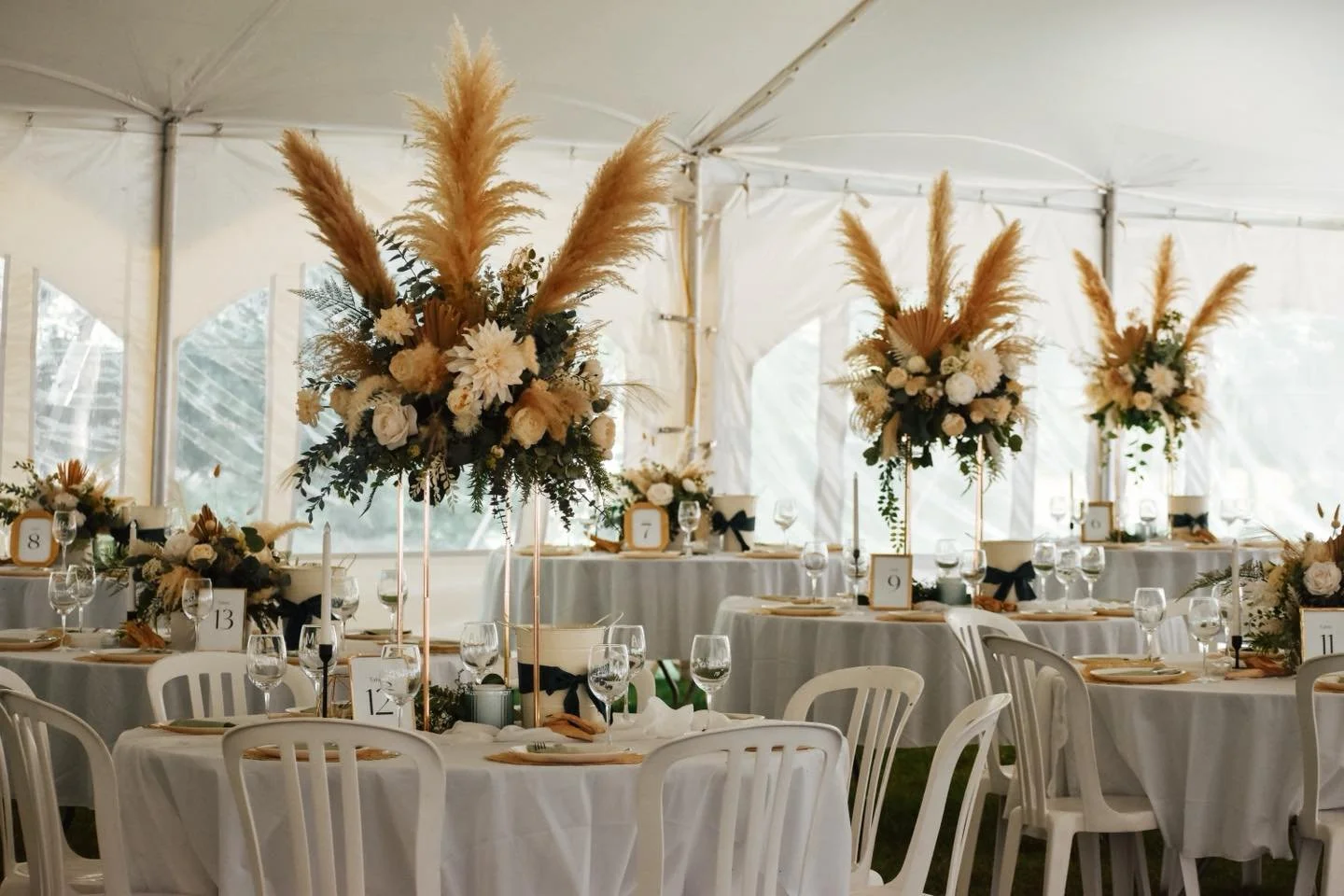 Décor de mariage avec des centres de table de fleurs blanches et pampas, tables rondes avec nappes blanches, vaisselle, verres et assiettes en lin beige, sous une tente blanche