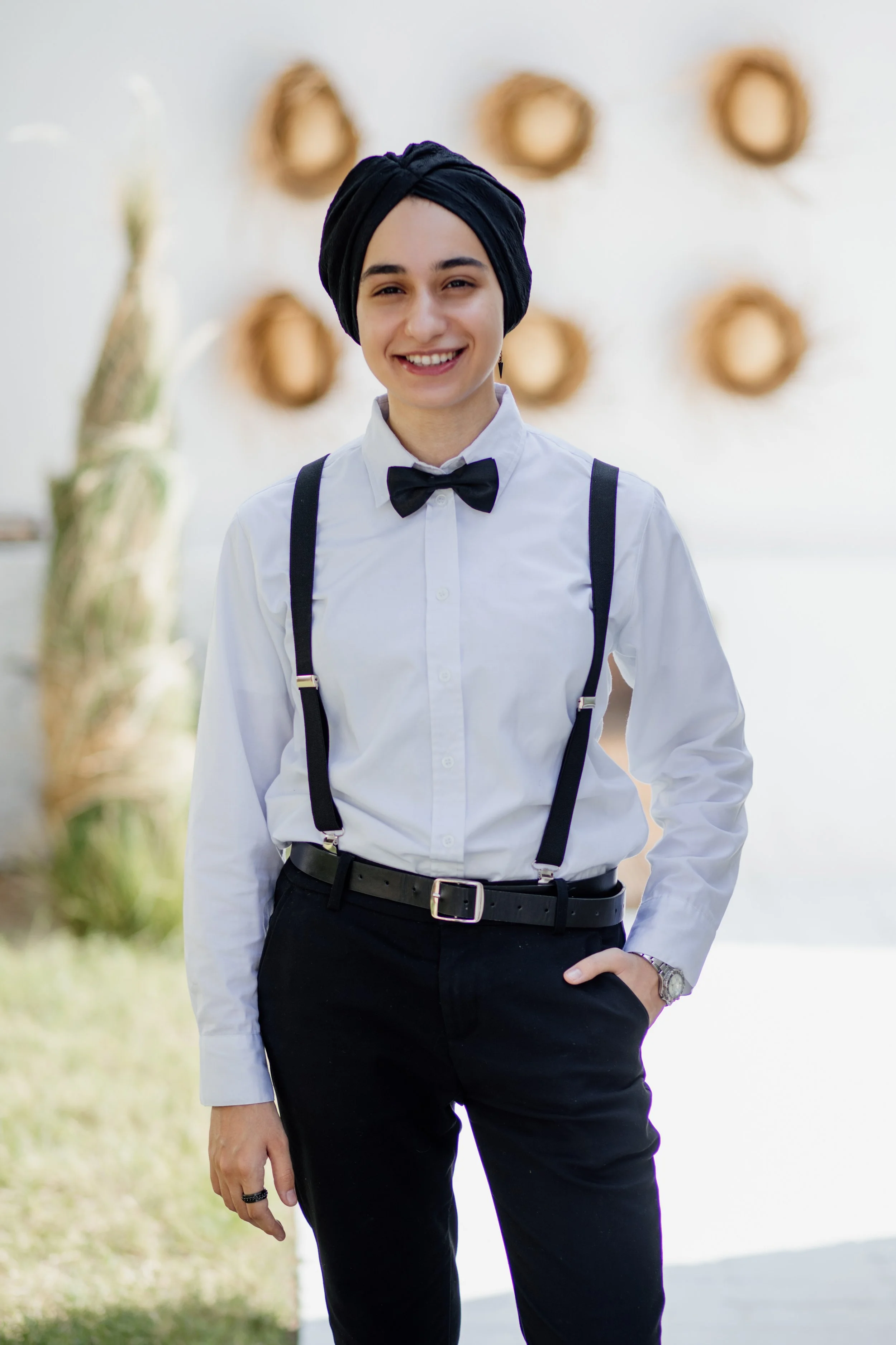 A person smiling, wearing a white shirt, black suspenders, and a black bow tie, with a black turban and black pants, standing outdoors with blurred round wreaths in the background.