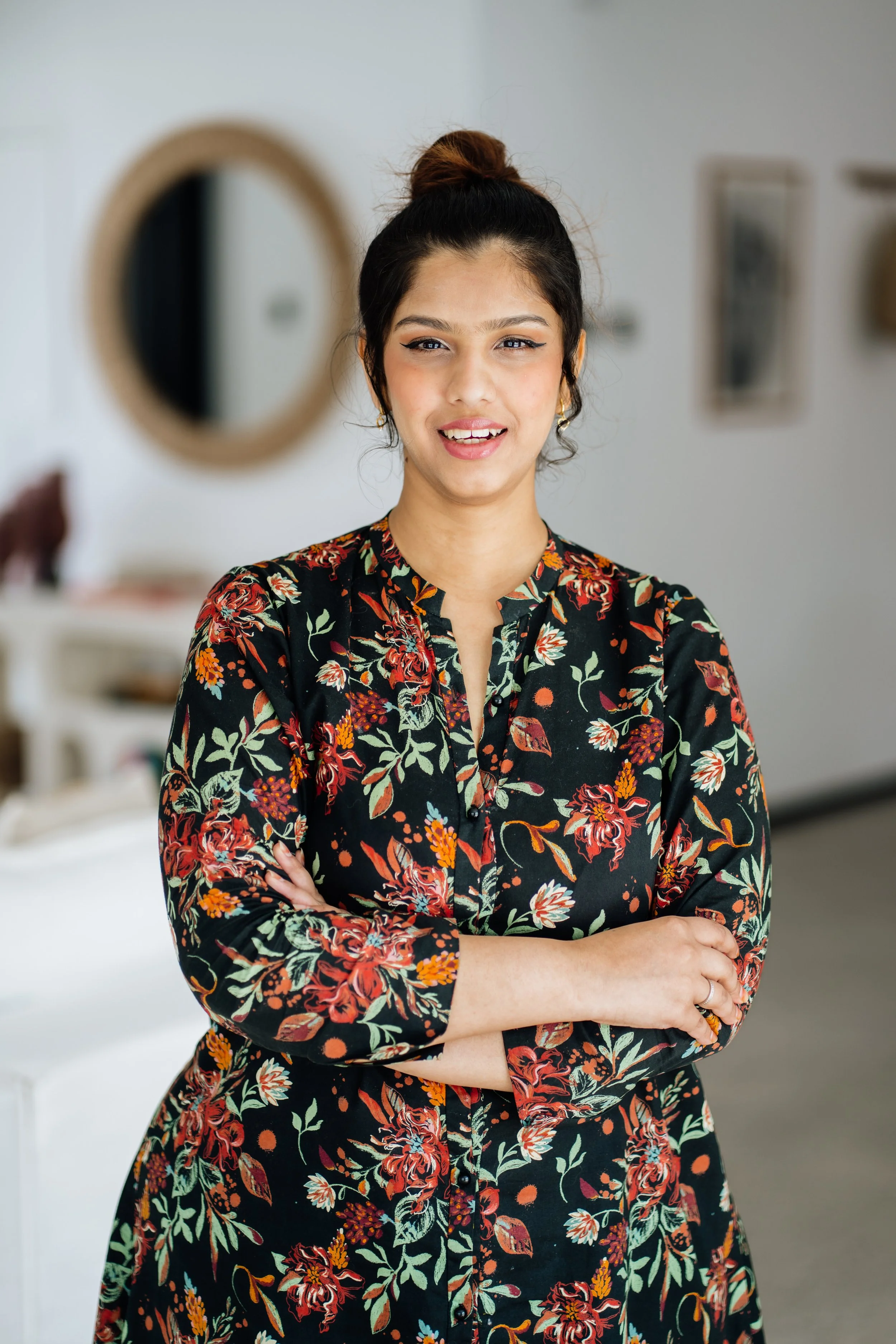 A woman with dark hair in a bun, wearing a floral dress, standing with her arms crossed, in a room with round mirror and framed pictures on the wall.
