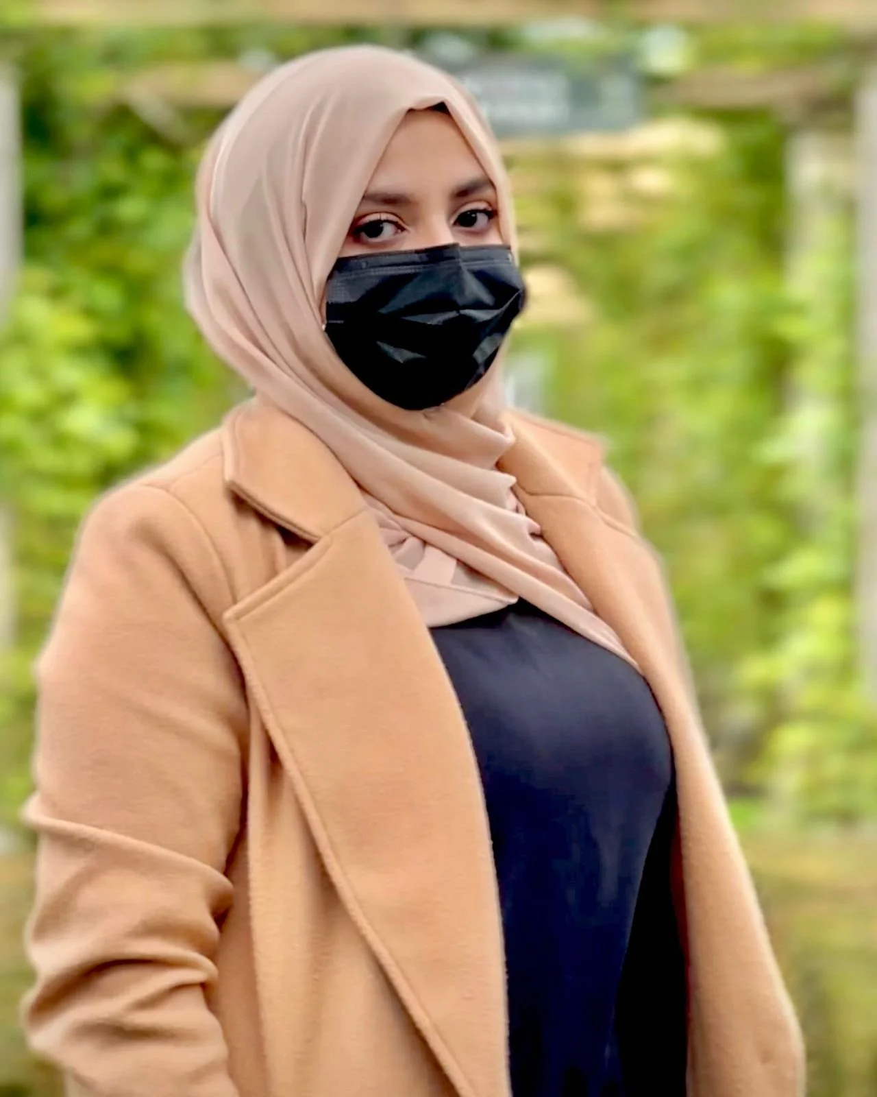 A woman wearing a beige coat and a tan hijab with a black face mask standing outdoors with a blurred green background.
