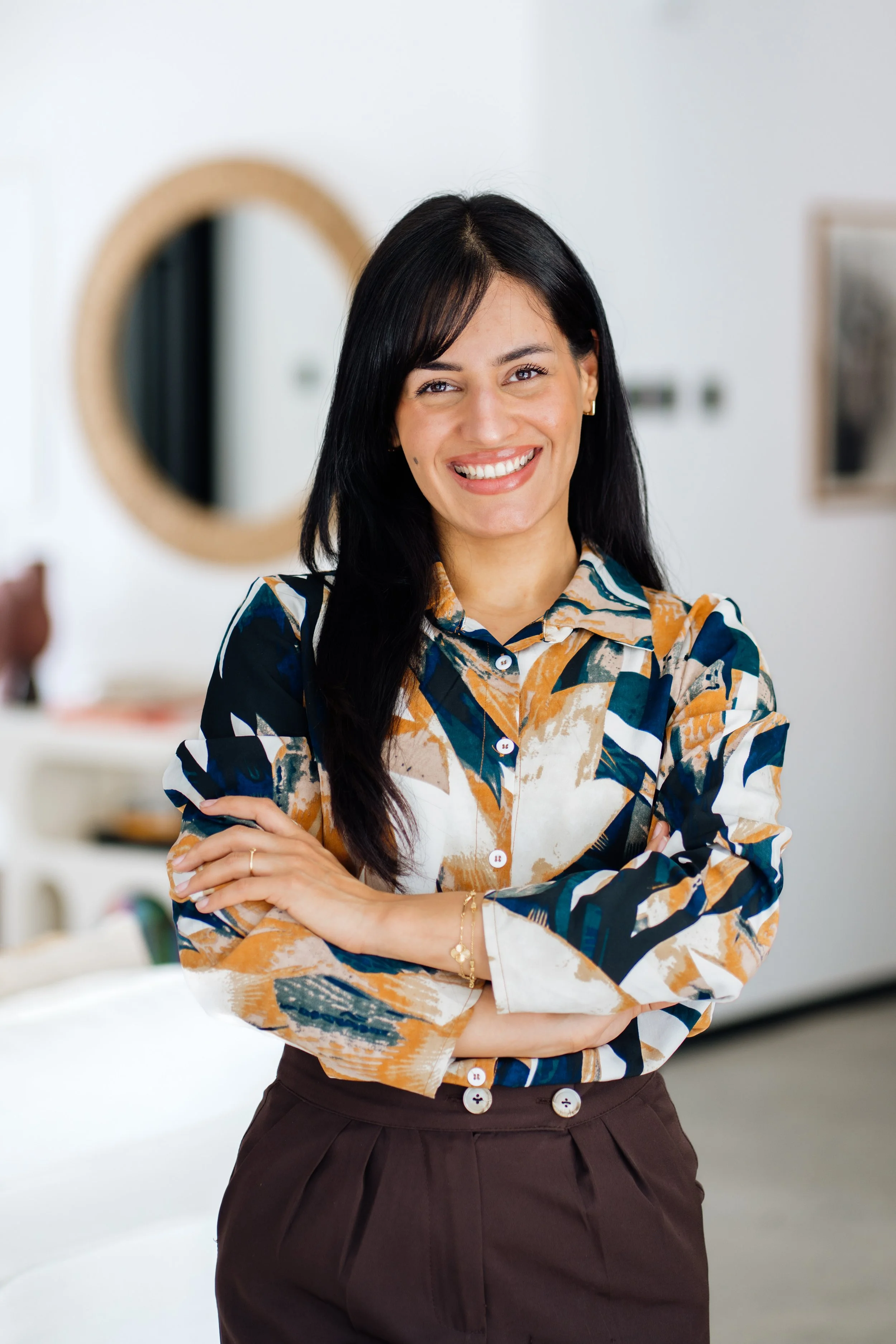 Smiling woman with dark hair, wearing a patterned blouse and dark pants, standing in a modern, well-lit room with a mirror and framed pictures.