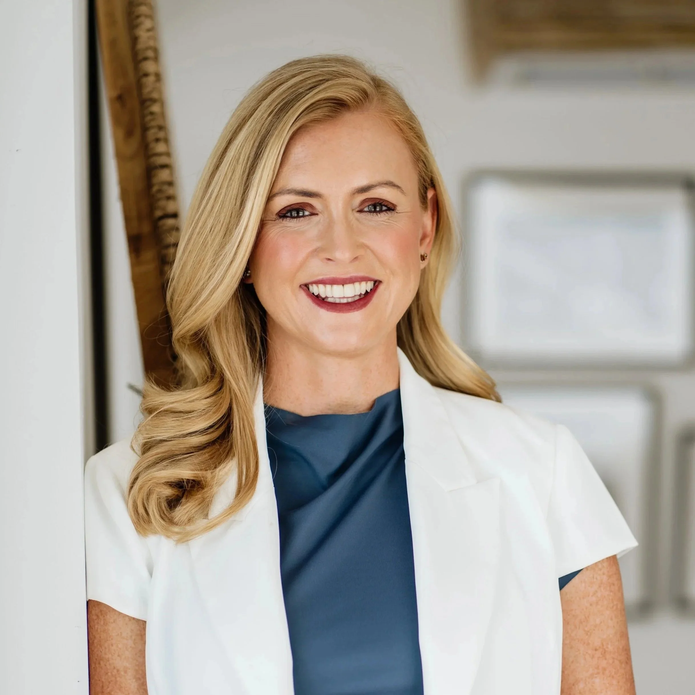A blonde woman with light skin, smiling, wearing a dark blue top and white blazer, standing indoors near a window with a blurred background.