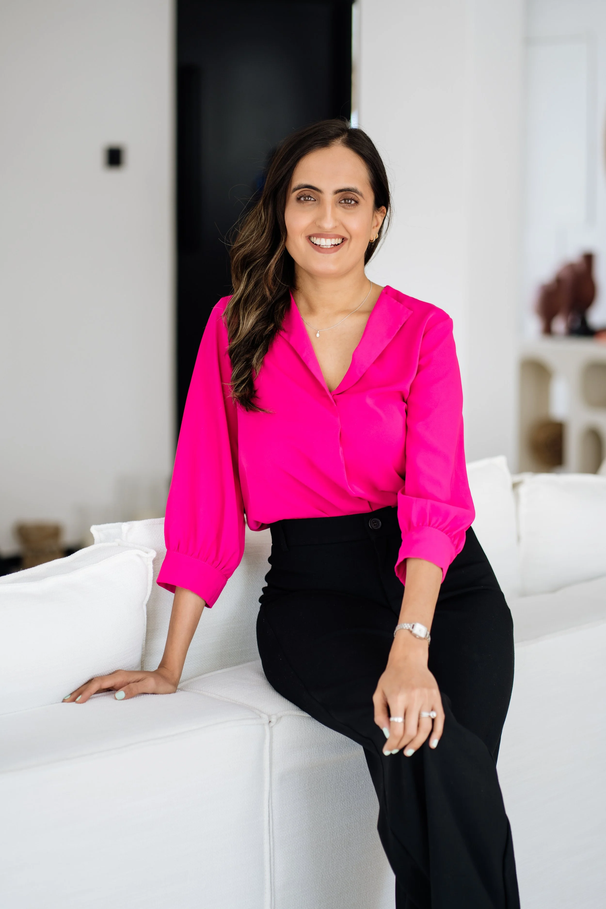 A woman with long dark hair, wearing a bright pink blouse and black pants, sitting on a white sofa in a modern, well-lit living room, smiling at the camera.