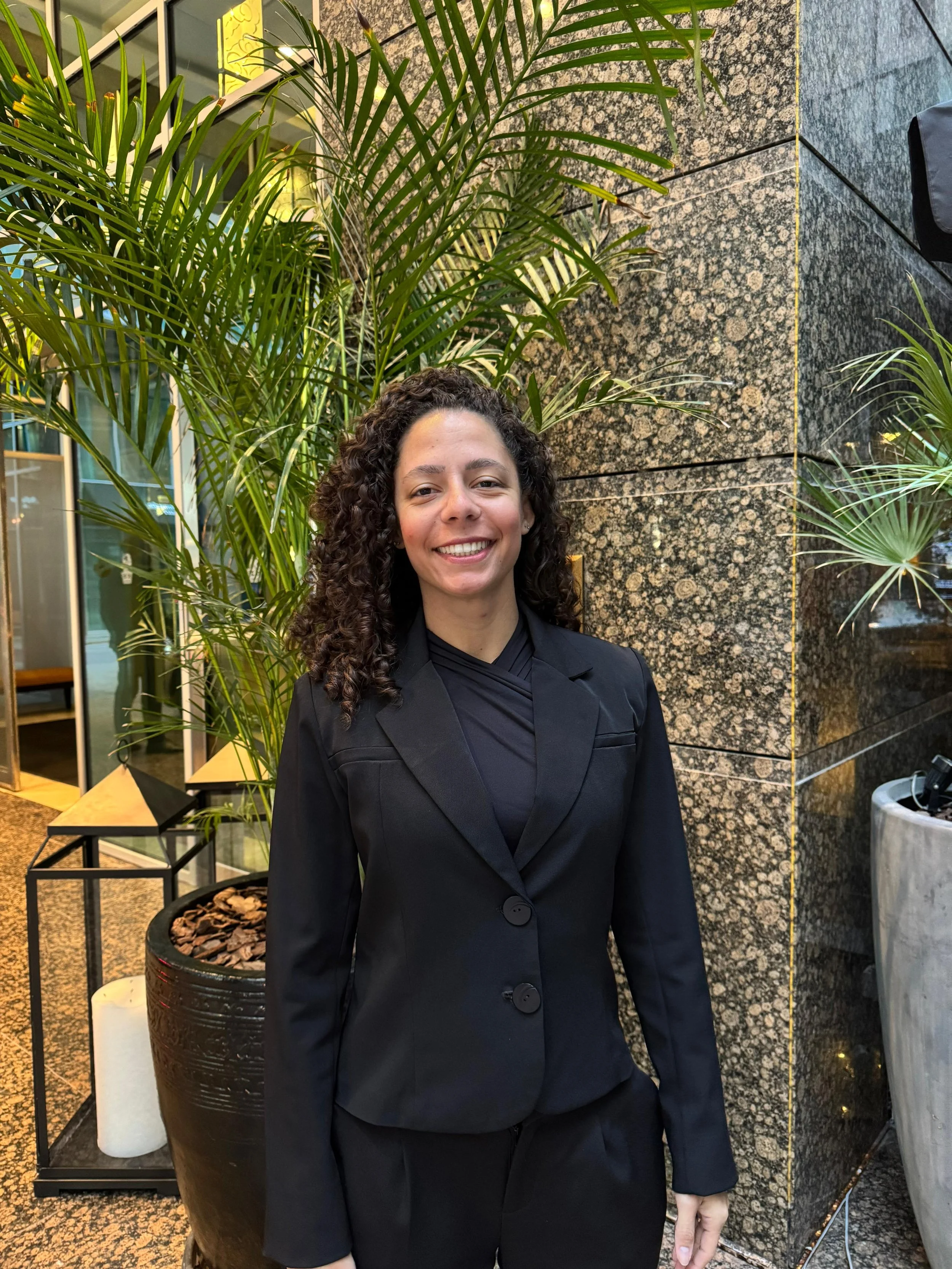 A woman with curly dark hair smiling, wearing a black blazer and matching black top, standing in front of green plants and a textured stone wall.