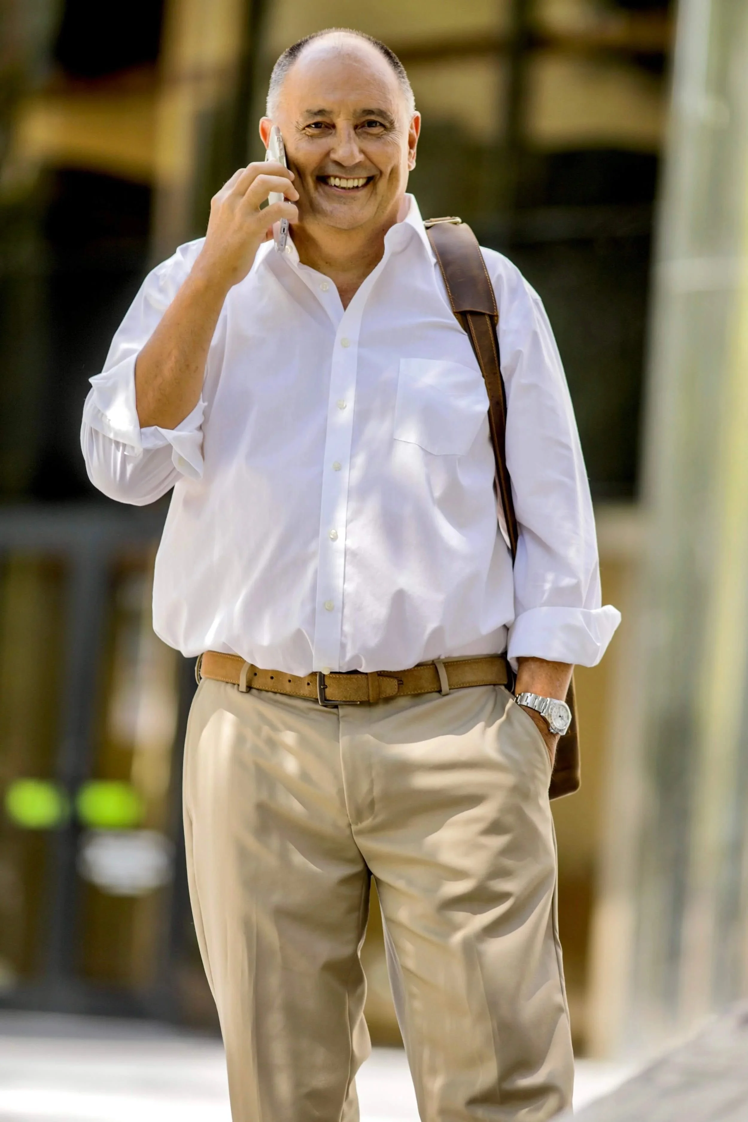 A man in a white shirt with a brown shoulder bag smiles while talking on the phone outdoors. The background features blurred glass and metal architecture, suggesting a casual yet professional moment of communication.