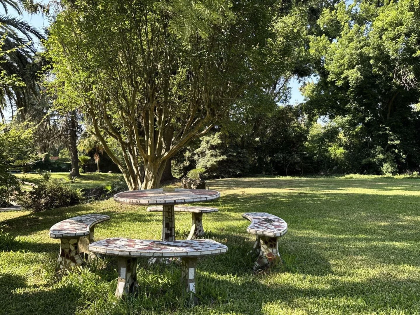 Shaded outdoor stone table and seating area beneath mature trees in a private park-like estate in Buenos Aires.