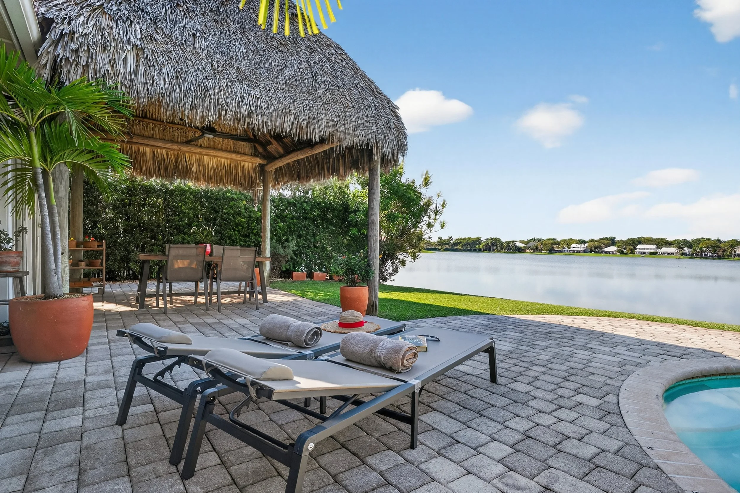 Lakeside outdoor lounge with palapa, sun loungers, and paved patio overlooking the water. Rolled towels and a red sunhat suggest relaxation, while a nearby dining table and lush greenery create an inviting space for entertaining.
