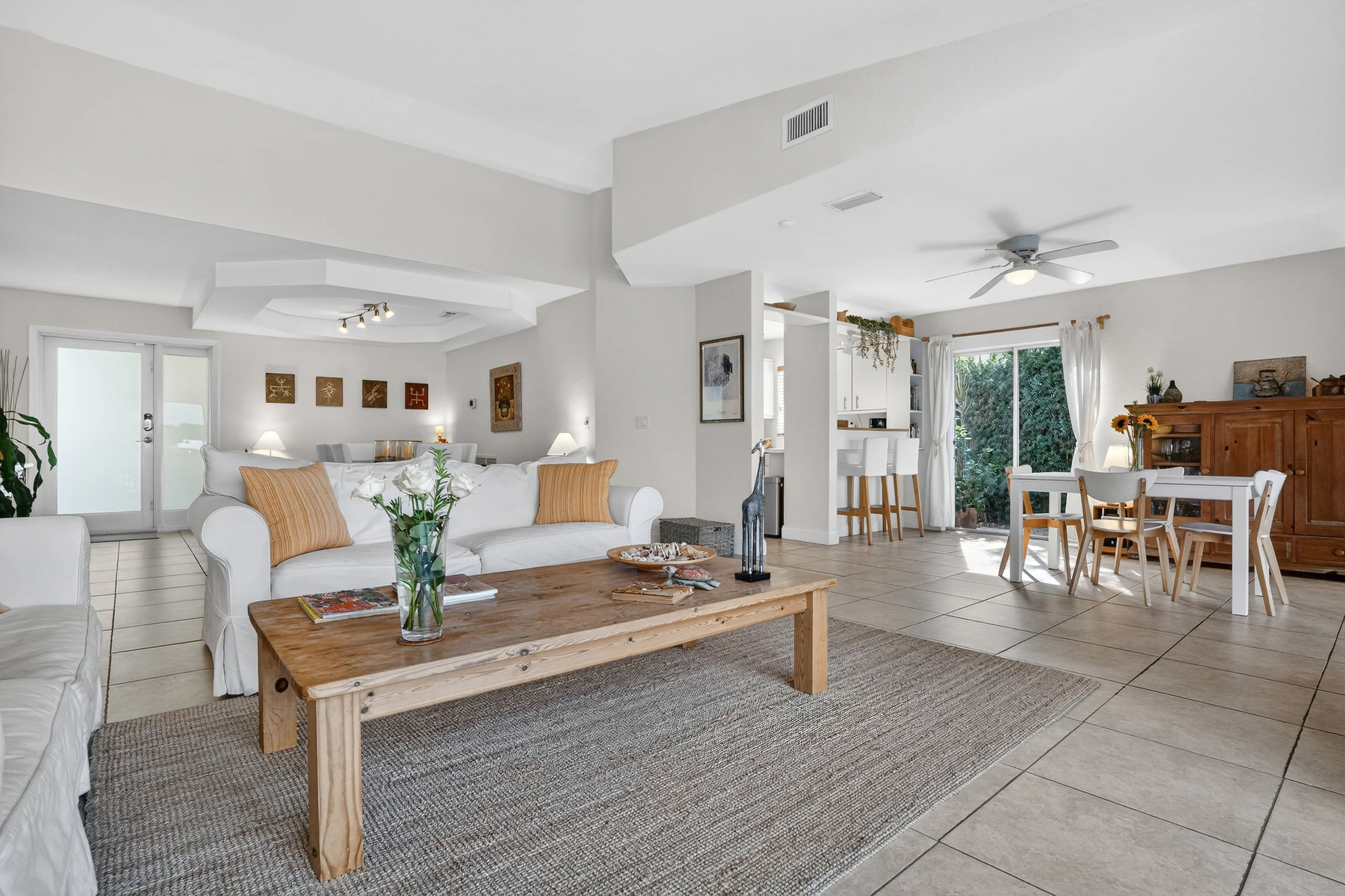 Spacious living room with neutral furnishings and an open layout connecting to the dining area. White sofa, wooden coffee table, and framed artwork create a cozy ambiance, with natural light streaming through large windows.