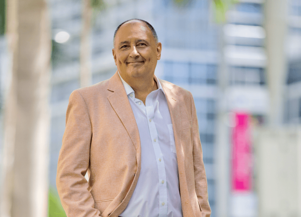 tylish person in a tan blazer and white shirt standing in a sunny urban setting with palm trees and modern buildings in the background.