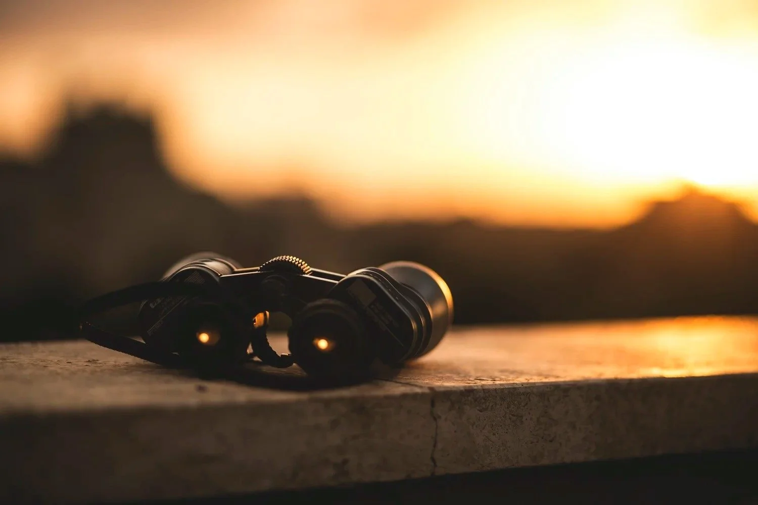A pair of black binoculars rests on a stone surface, facing the viewer. Behind them, a warm sunset casts a golden glow over a blurred landscape, evoking a peaceful, contemplative mood.