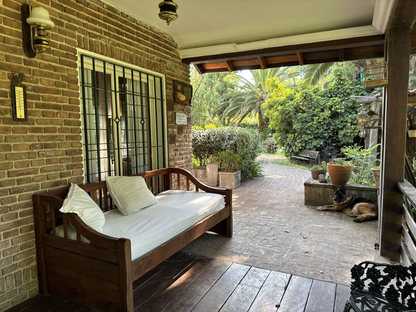 Covered brick gallery with wooden daybed and garden views, connecting indoor and outdoor living spaces.