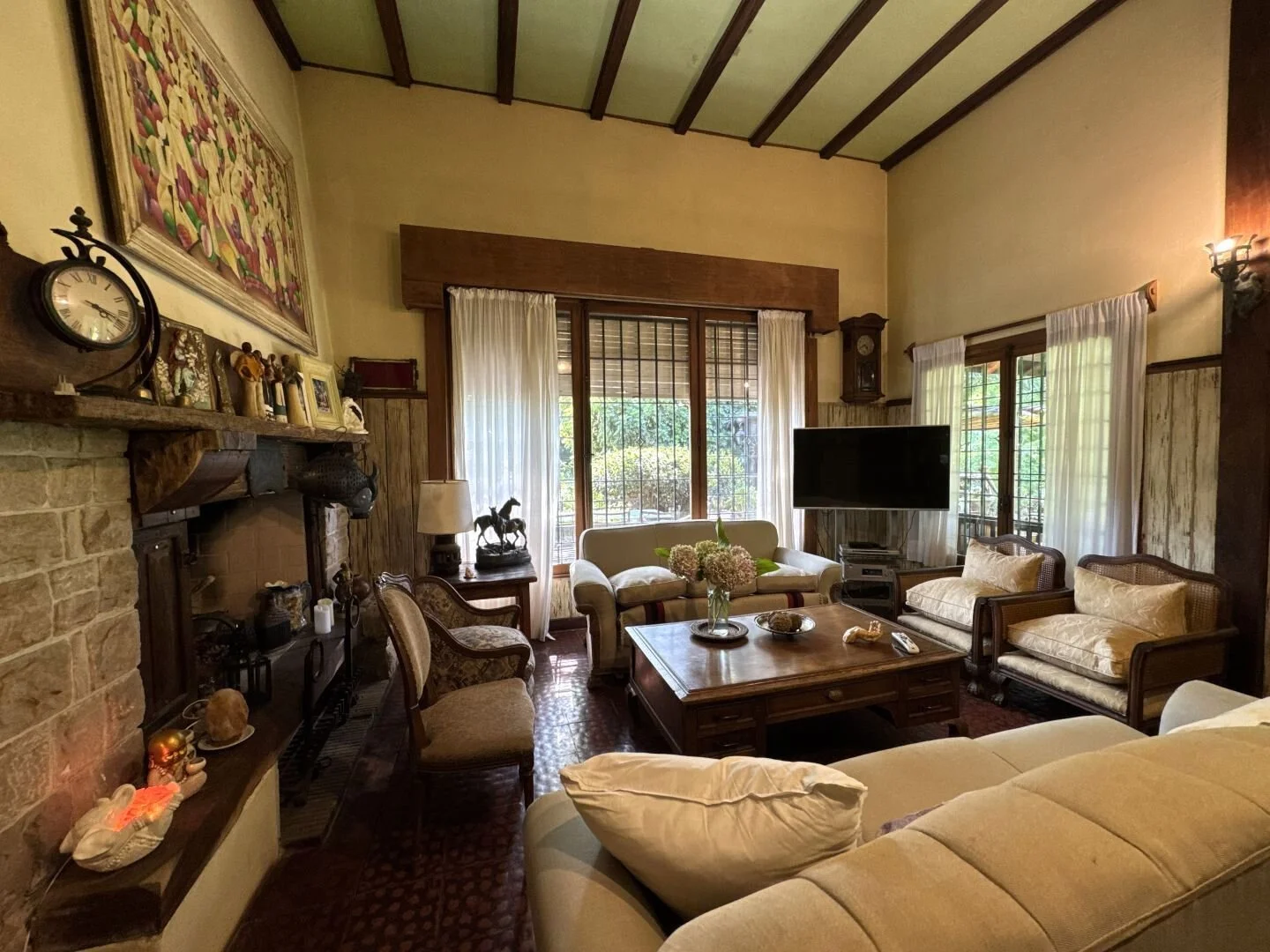 Traditional living room with exposed wood beams, brick fireplace, large windows overlooking the garden, and classic wood furnishings.