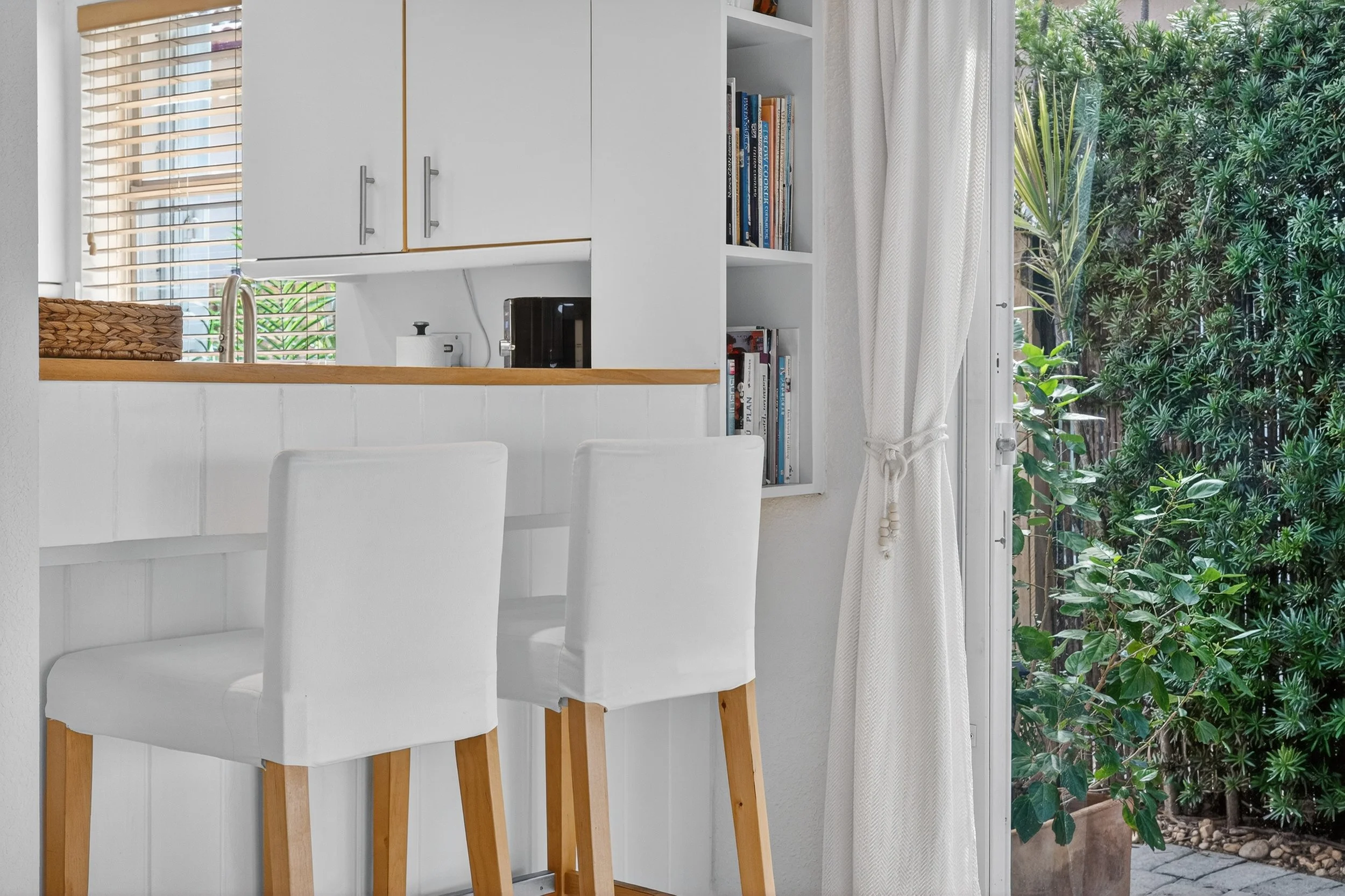Bright kitchen breakfast bar with white cabinetry, wooden countertop, and upholstered stools. Large glass doors open to a lush garden patio, blending indoor comfort with vibrant outdoor views.