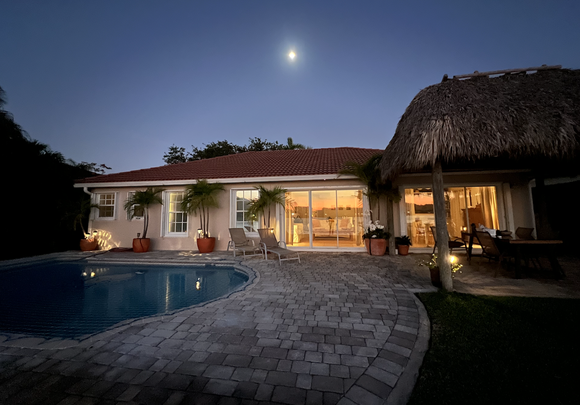 Night view of a lakeside home in Cutler Bay with illuminated interiors, private pool, and tiki hut. The house overlooks calm water under a full moon, creating a serene and inviting tropical atmosphere.
