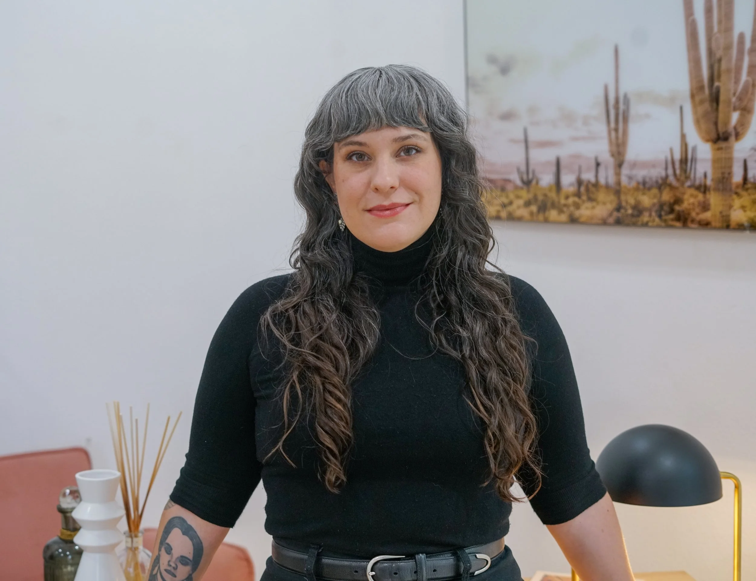 A compassionate grief and trauma therapist with wavy, shoulder-length gray hair, wearing a white blouse and a silver necklace, against a gray background — representing trauma-informed, attachment-based therapy for individuals, couples, and families.