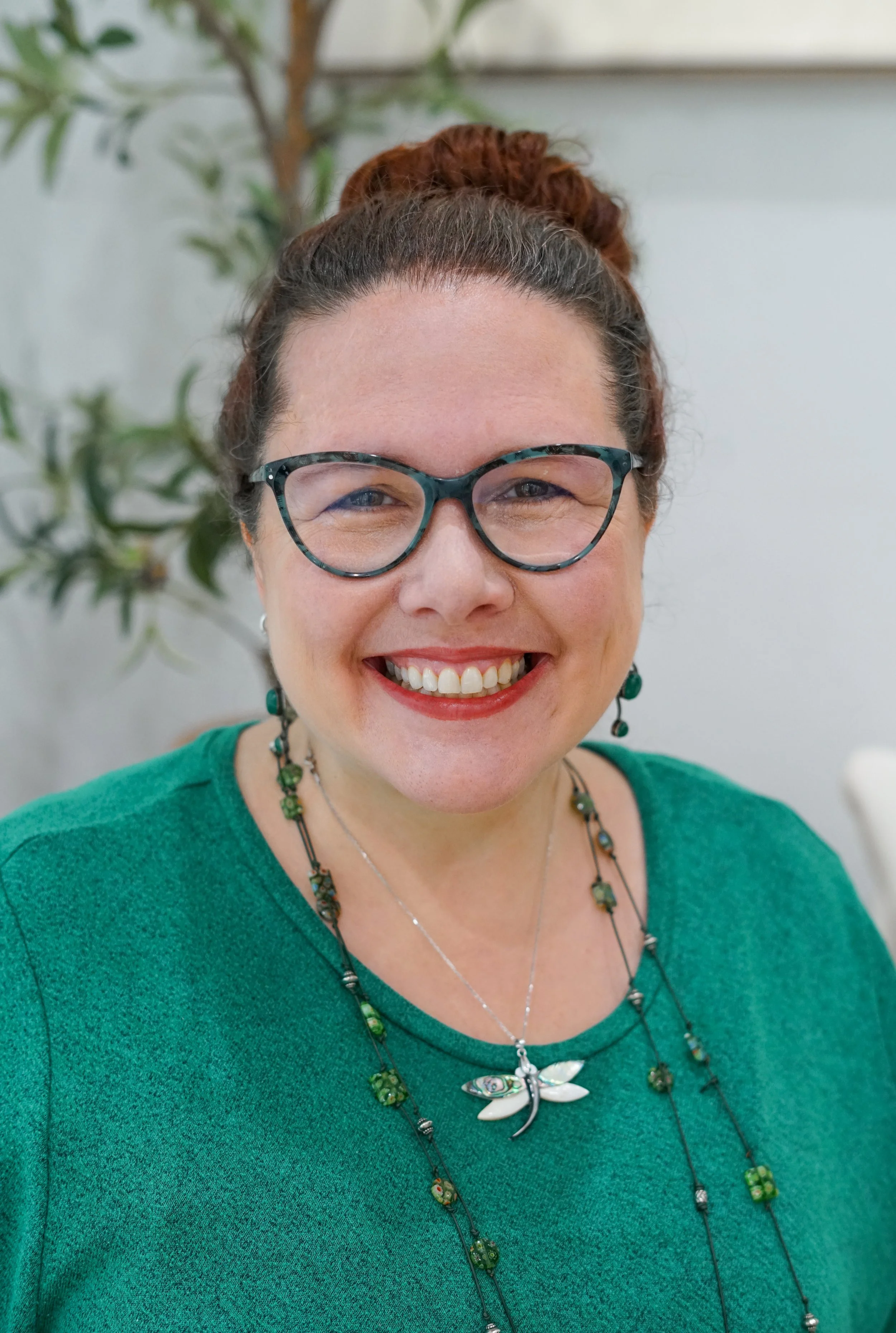 A woman with shoulder-length brown hair and brown eyes, wearing red lipstick, a black blazer, and silver earrings, against a black background.