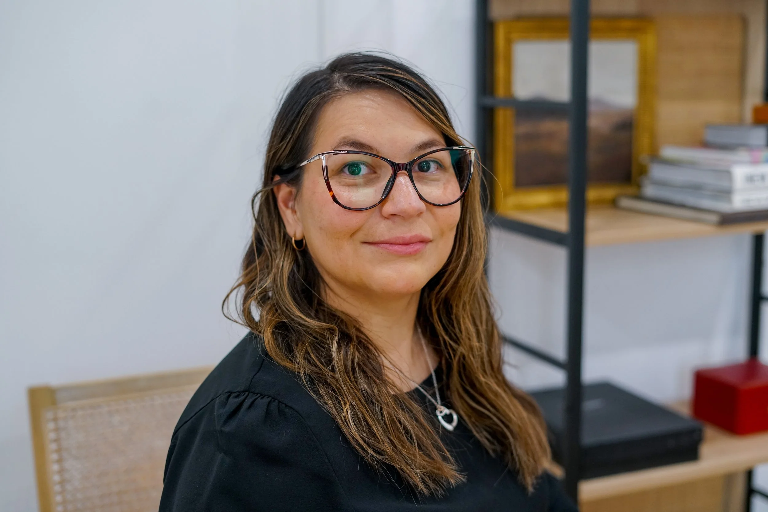 A woman with long wavy brown hair, glasses, and hoop earrings smiling in front of a plain background.