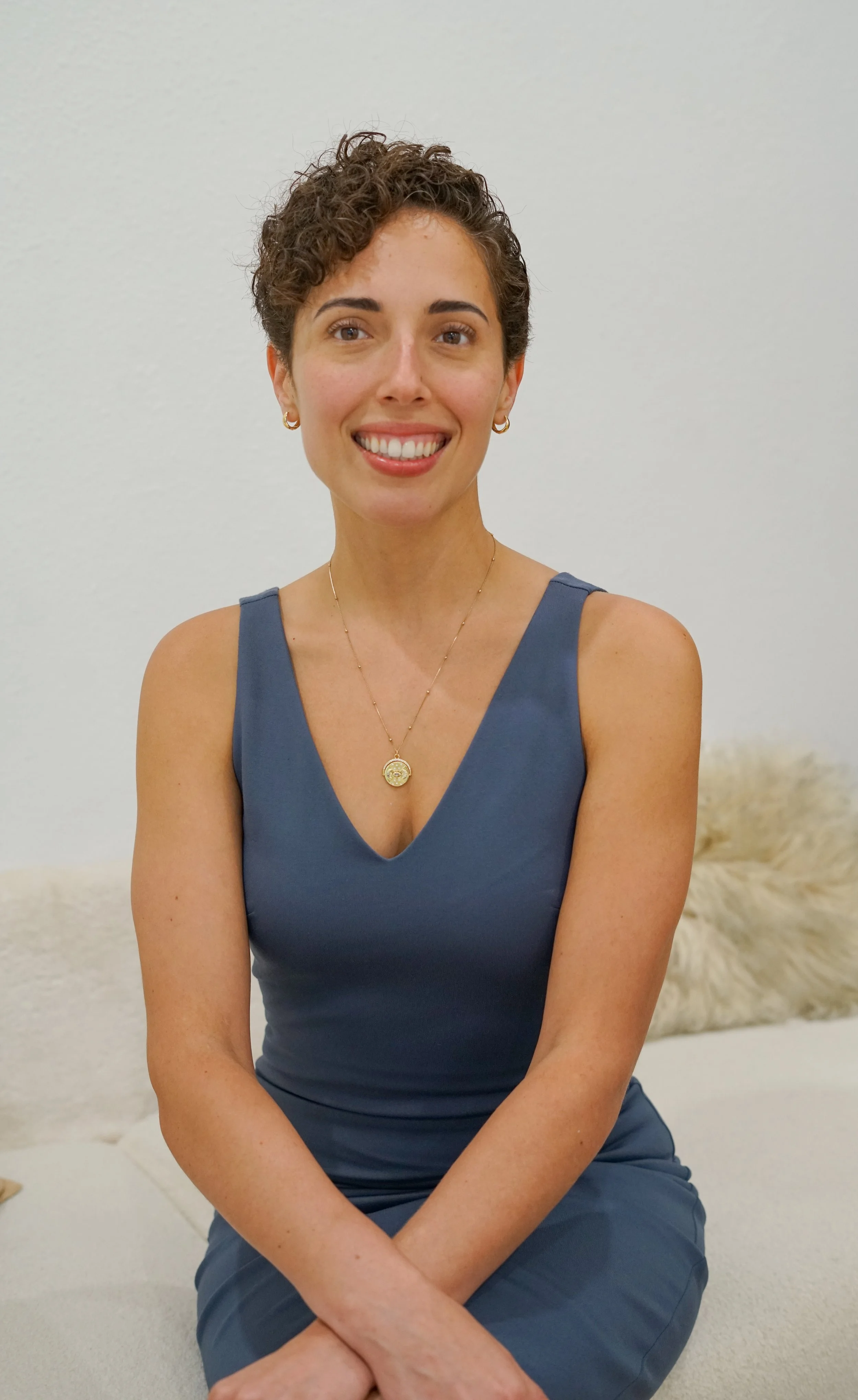 Portrait of a woman with long, curly brown hair, light makeup, and a smile, wearing a black blazer against a dark background.