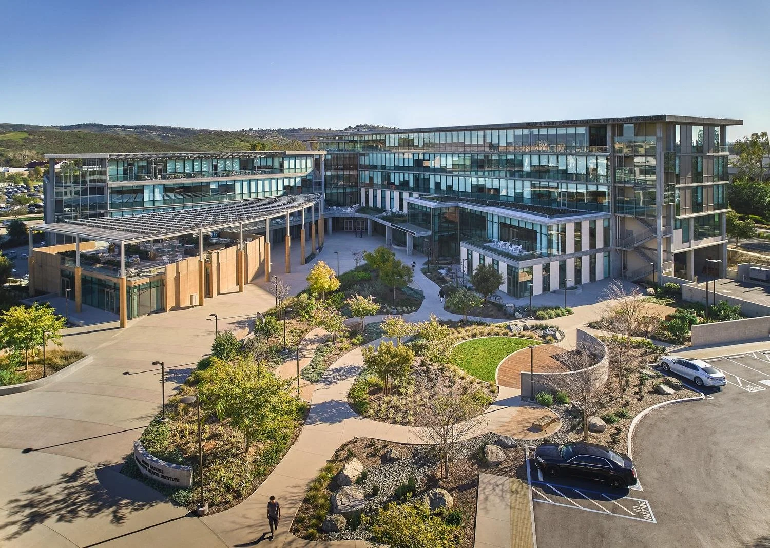 A modern multi-story educational or medical building with glass windows, surrounded by landscaped walkways, trees, and parking lots on a clear day.