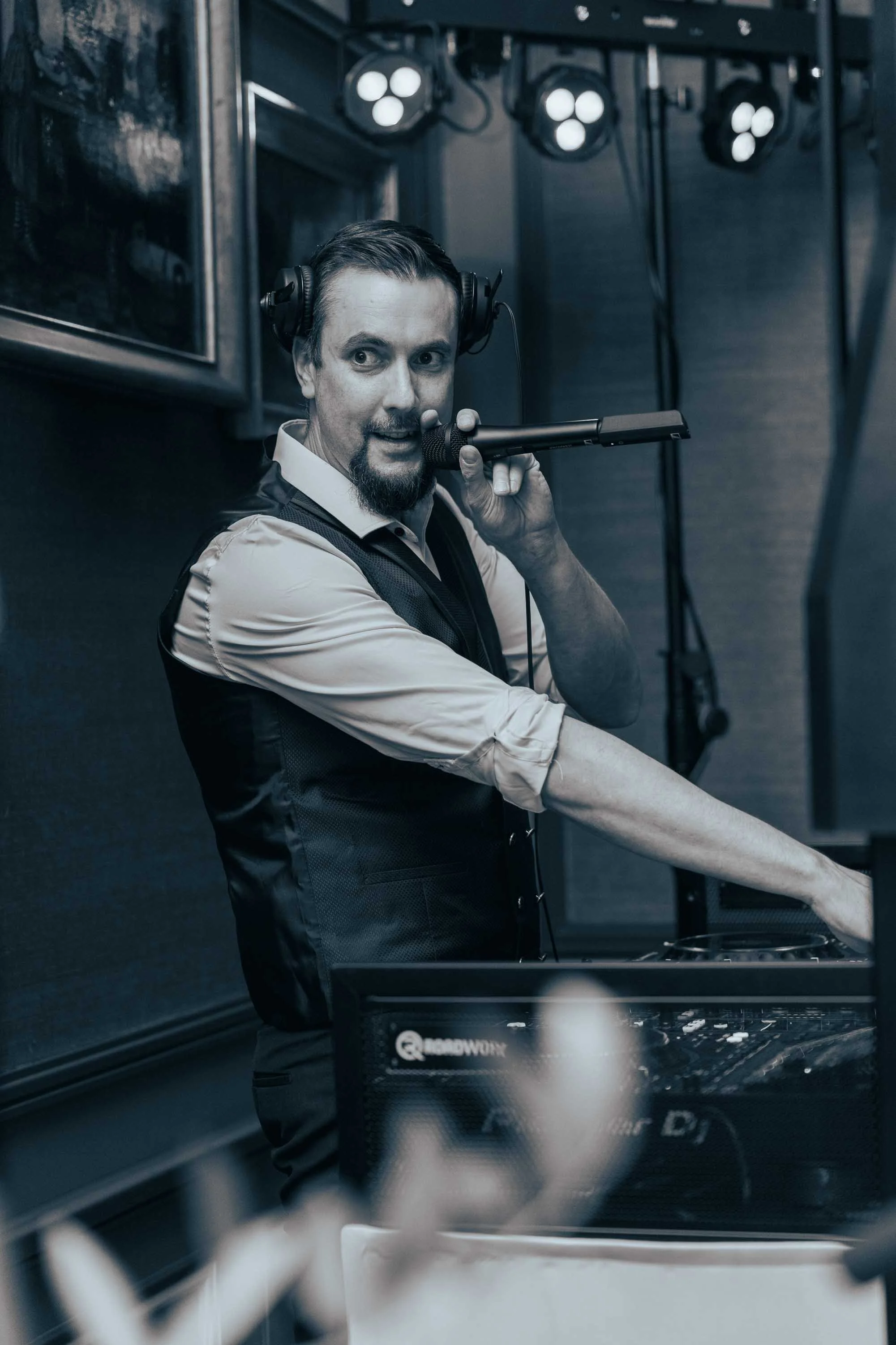 Mann beim DJ-Auflegen, trägt Kopfhörer, hält Mikrofon, in dunklem Raum mit Bühnenlichtern.