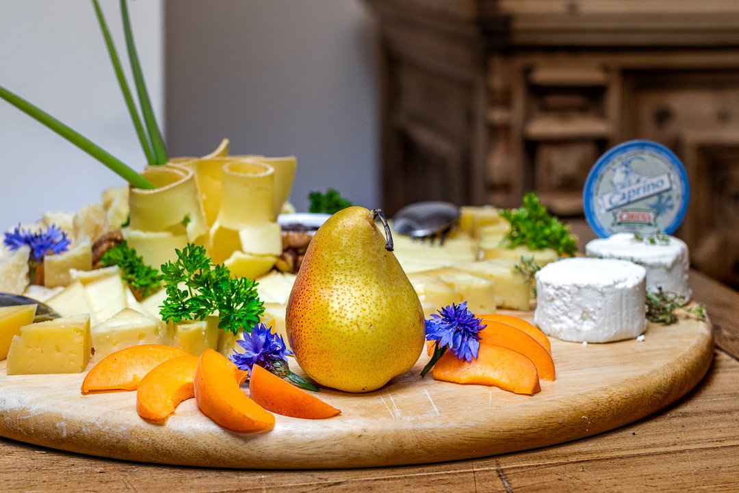 Käseplatte mit verschiedenen Käsesorten, Birne, Aprikosenscheiben, blauen Blumen und Kräutern, auf einem runden Holzbrett arrangiert.