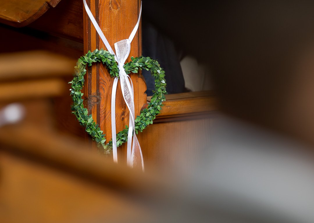 Herzförmiger Kranz aus grünen Blättern mit weißem Band an einer Holzoberfläche.