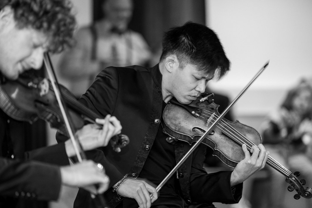 Zwei Musiker spielen konzentriert auf Streichinstrumenten, vermutlich Violinen, in einem Innenraum. Die Aufnahme ist schwarz-weiß. Der Fokus liegt auf einem Mann rechts im Bild.