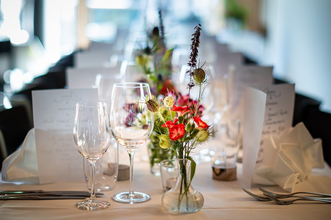 Elegante Tischdekoration mit Weingläsern, Servietten und einem kleinen Blumenstrauß in einer Glasvase auf einem gedeckten Esstisch.