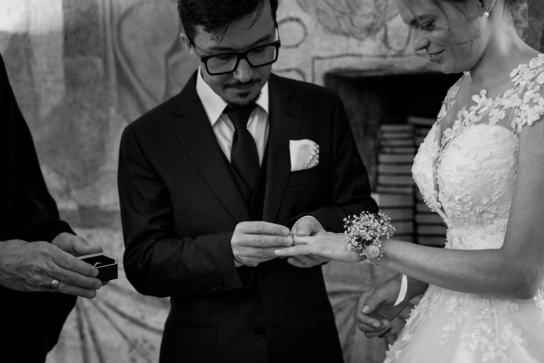 Ein Bräutigam steckt der Braut bei einer Hochzeit einen Ring an den Finger. Beide sind elegant gekleidet und lächeln. Im Hintergrund sind Bücher sichtbar.