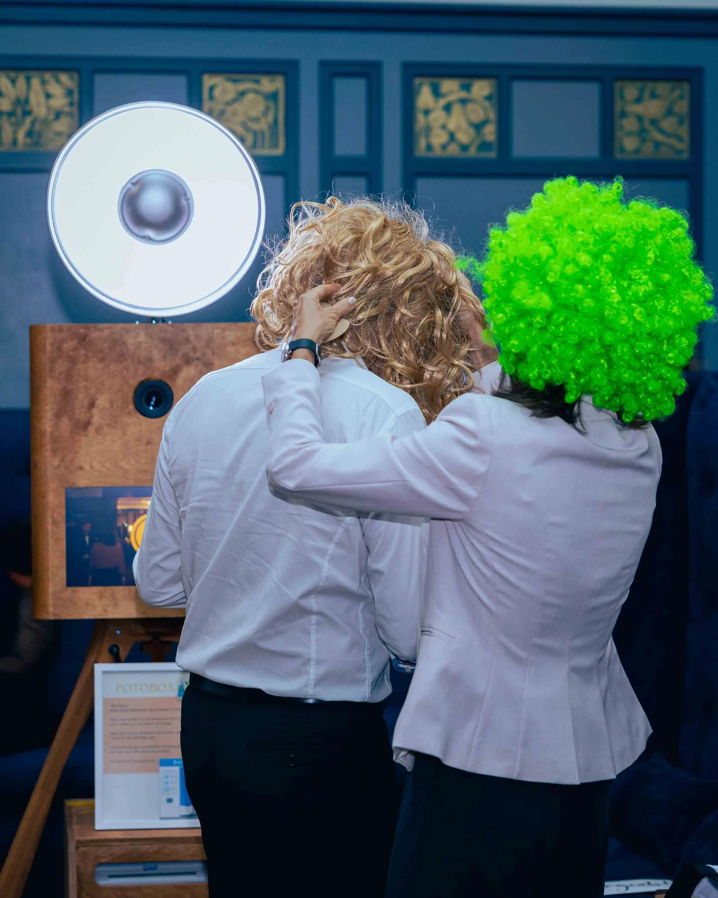 Zwei Frauen in weißen Blusen, eine mit roten, lockigen Haaren und die andere mit einem grellen grünen Perückchen, umarmen sich auf einer Messe oder Veranstaltung, im Hintergrund ein Lautsprecher und eine Fotobox.