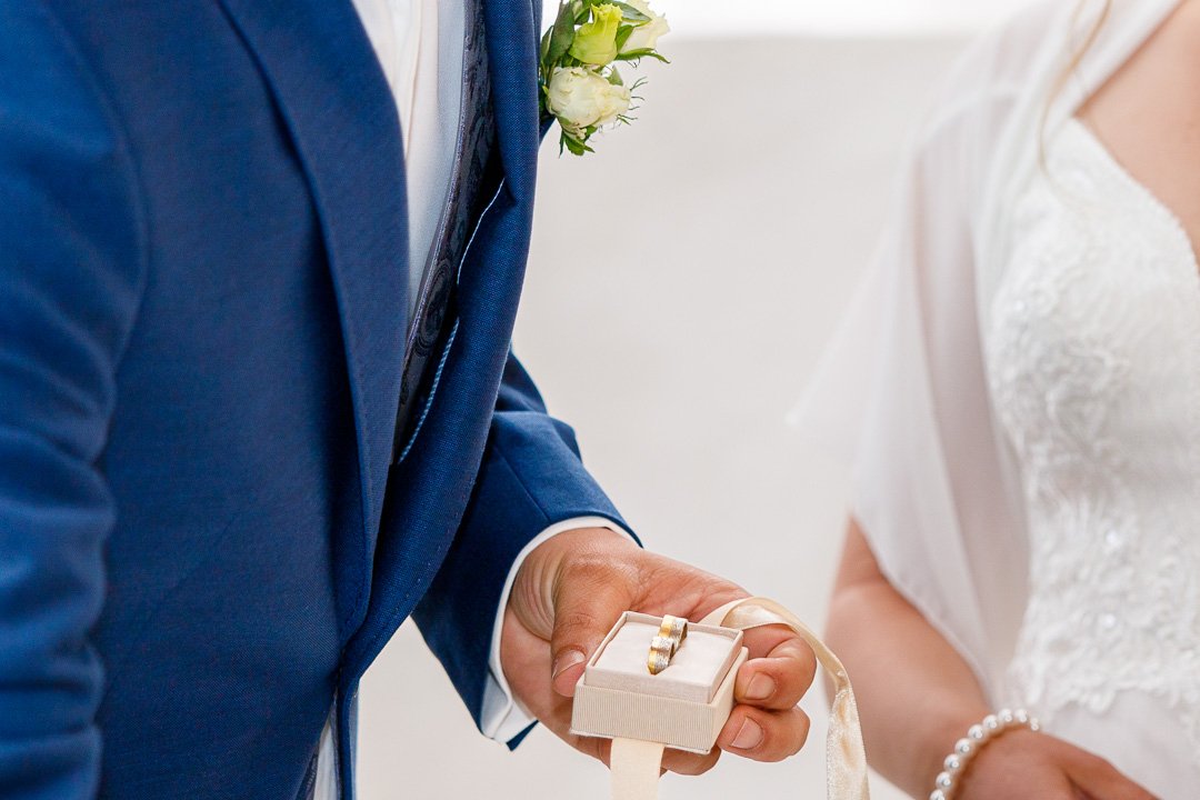 Mann im blauen Anzug hält Ringbox mit einem goldenen Ring neben einer Frau im Brautkleid.