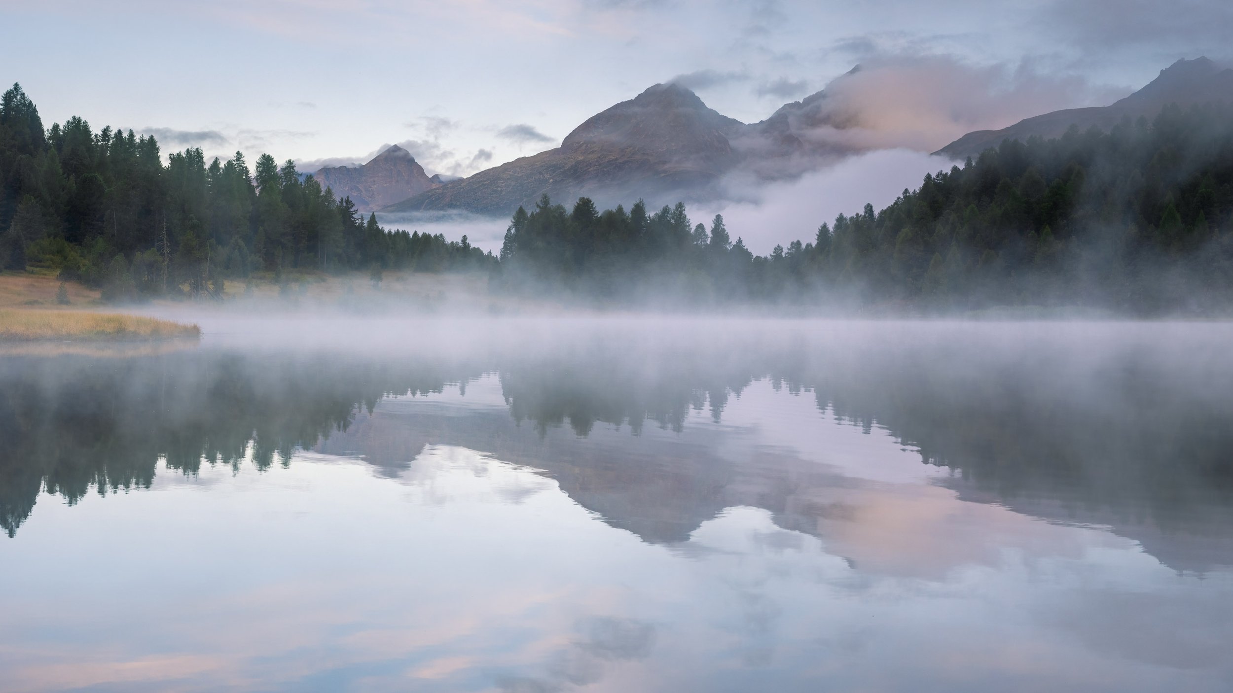 Nebel über dem Stazersee mit Spiegelung der Engadiner Bergwelt in den frühen Morgenstunden.