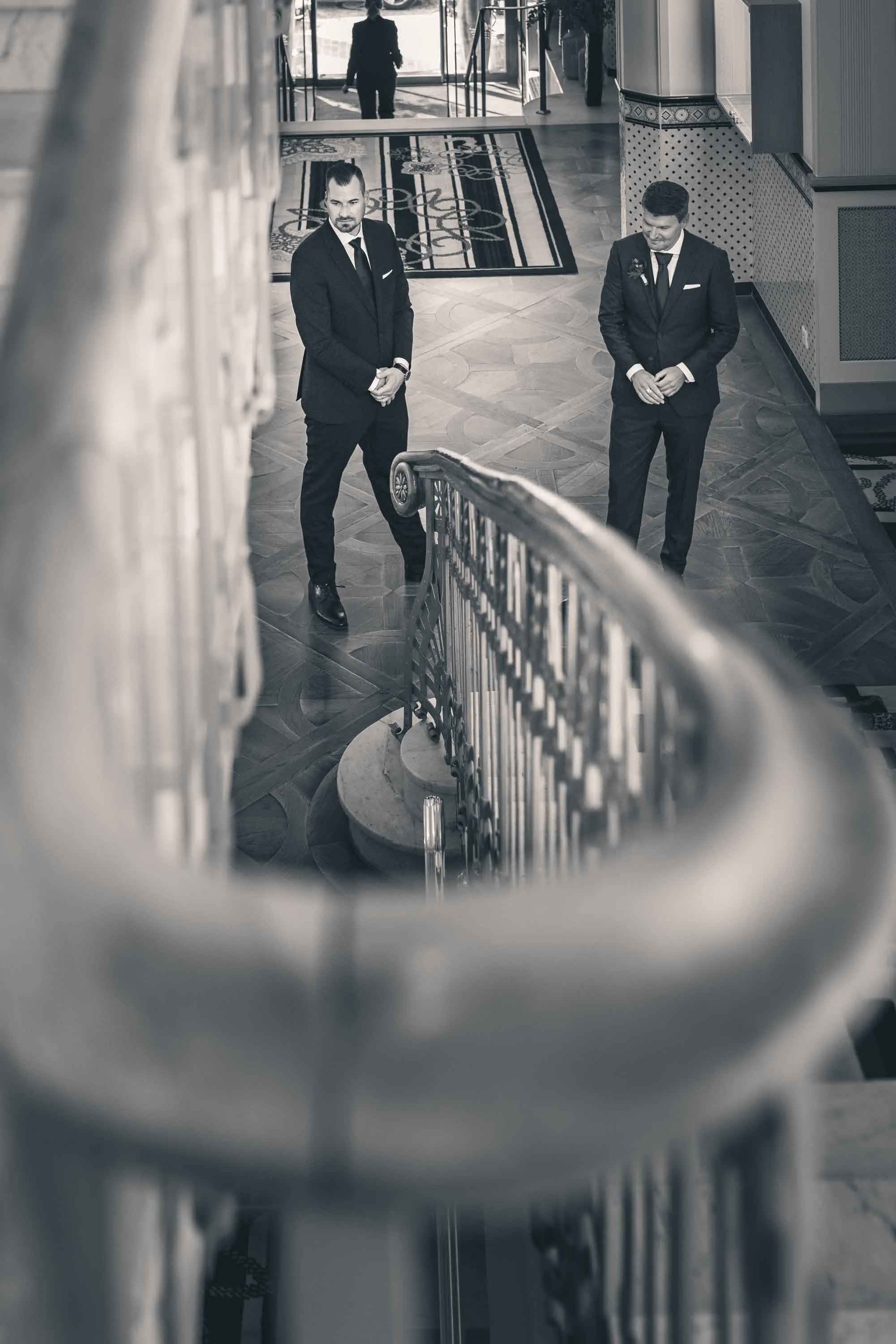 Zwei Männer in schwarzen Anzügen stehen auf einer Treppe in einem eleganten Gebäude, gesehen von oben durch eine Geländeröffnung.