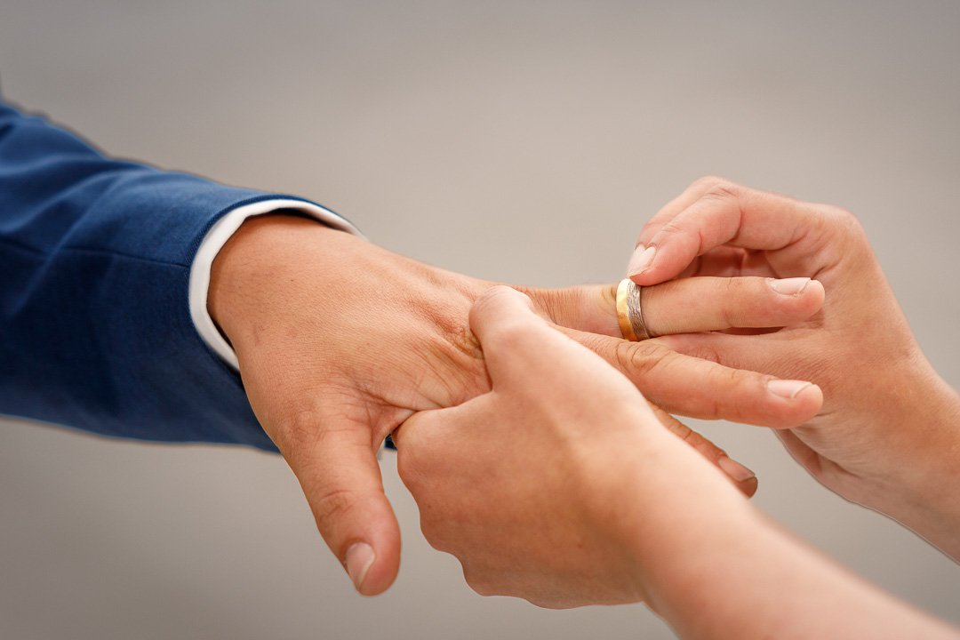 Nahaufnahme einer Person, die einem anderen eine Ehering an den Finger steckt, symbolisiert eine Hochzeit oder Verlobung. Hände in formeller Kleidung.