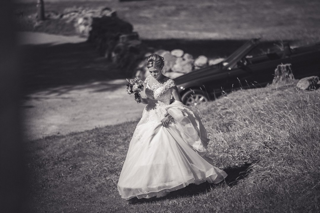 Eine Braut in einem eleganten Hochzeitskleid hält einen Blumenstrauß und geht im Freien einen grasbewachsenen Hang hinauf. Im Hintergrund steht ein geparktes Auto.