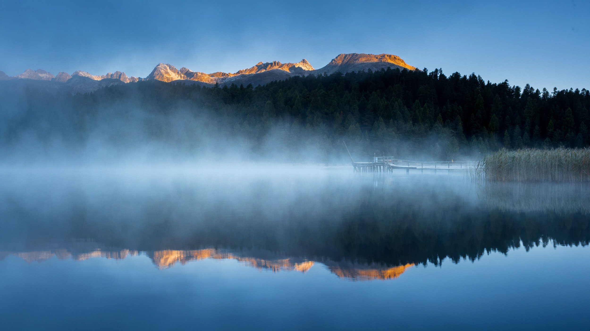 Morgennebel über dem Stazersee mit Spiegelung der sonnenbeschienenen Berggipfel.
