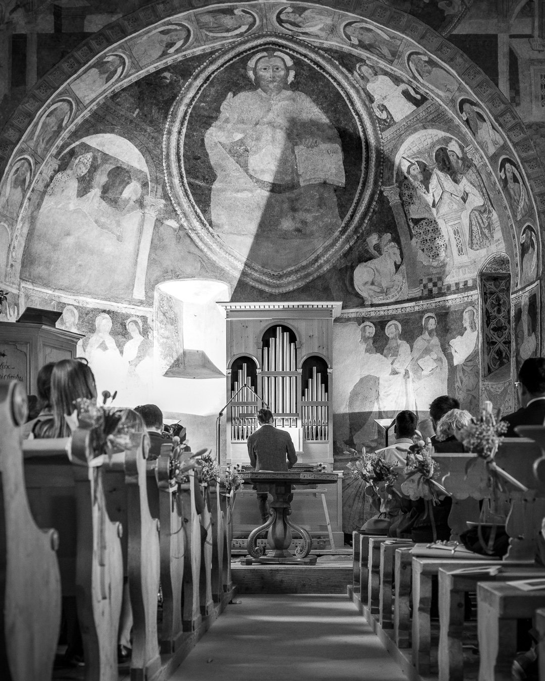 Schwarz-weiß Bild einer Kircheninnenansicht mit Fresken an der Wand, einem Organisten an einer Orgel, und Menschen in Holzbänken, die zuhören.