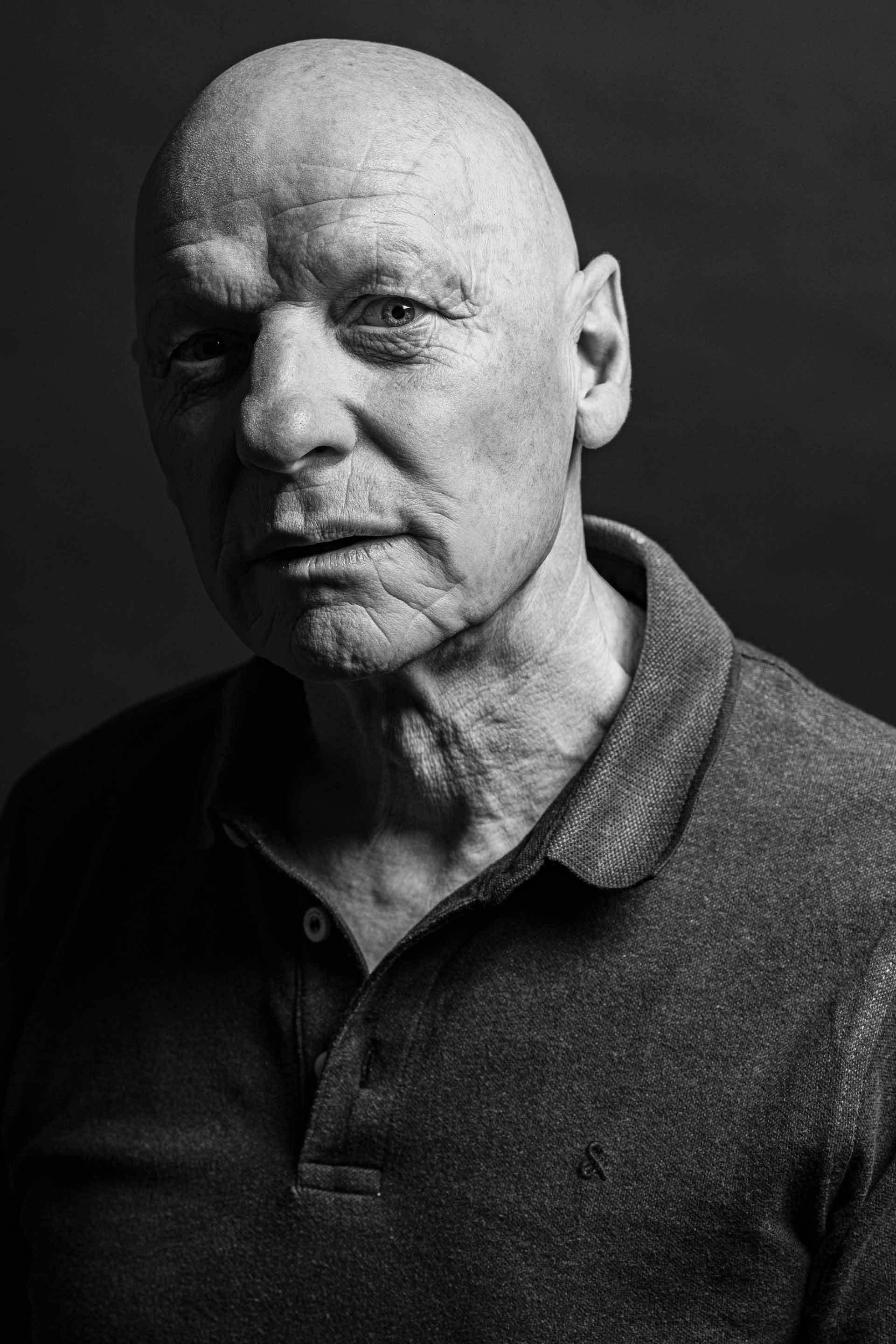 Schwarz-weiß Portrait eines älteren Mannes mit faltiger Haut, kurzem Haar, in einem dunklen Polohemd, frontal blickend mit nachdenklichem Ausdruck.