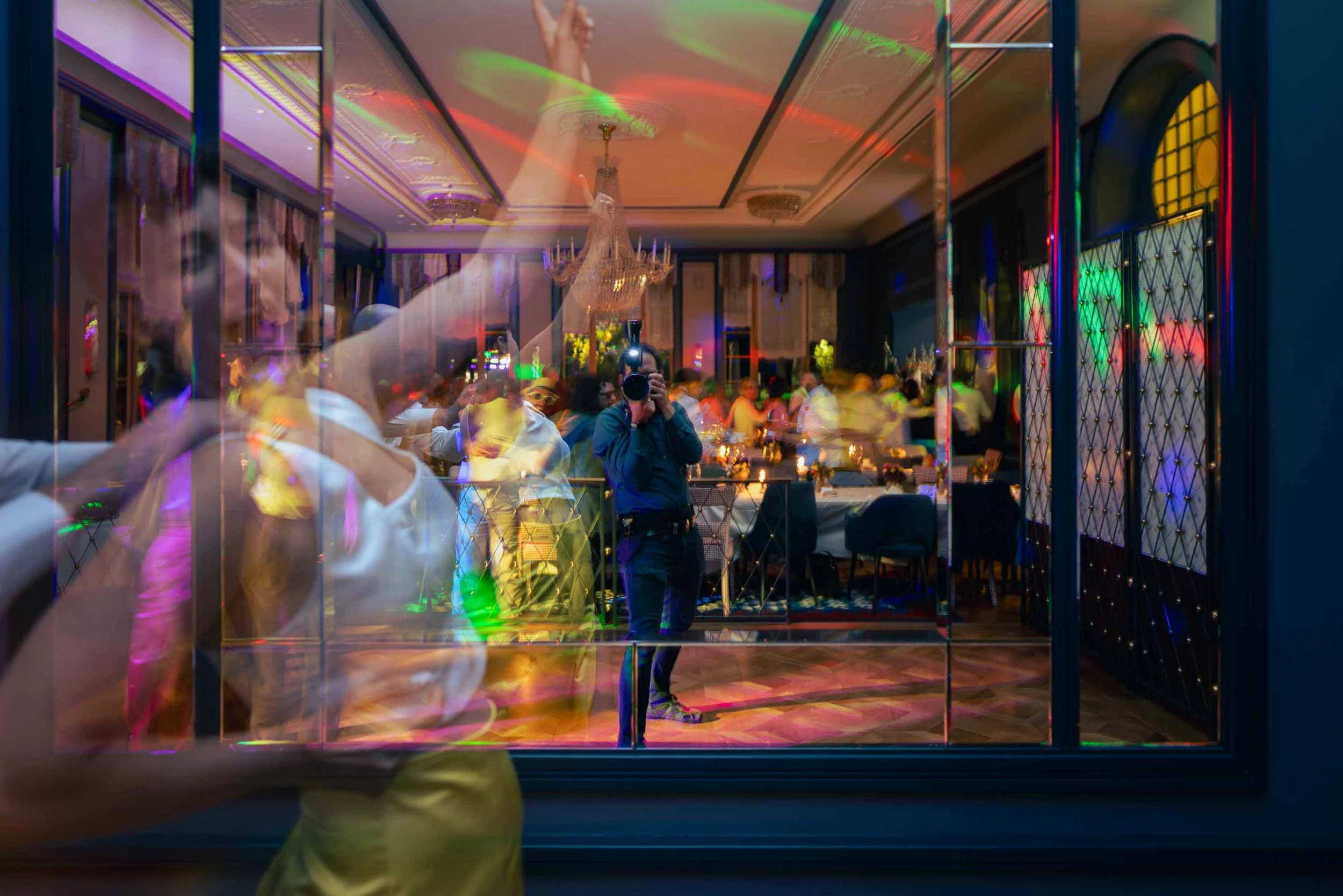 Blick durch eine Fensterscheibe auf eine festlich gedeckte Ball- oder Partyveranstaltung mit mehreren Menschen, Kerzen und bunten Lichtreflexen, während im Vordergrund eine Person ein Foto macht.