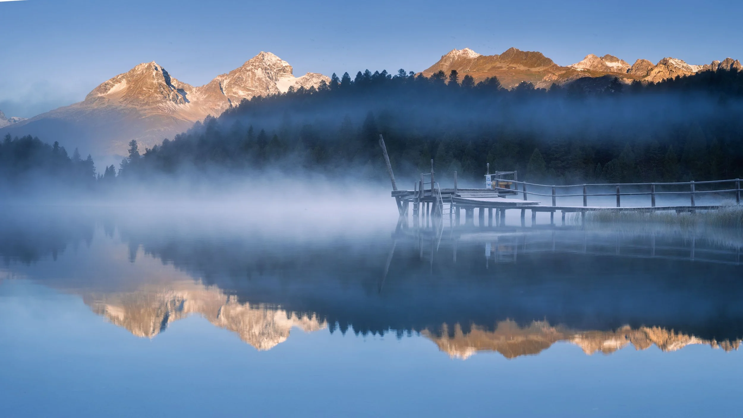 Historischer Badesteg am Stazersee mit Nebel und Bergspiegelung im Herbst.