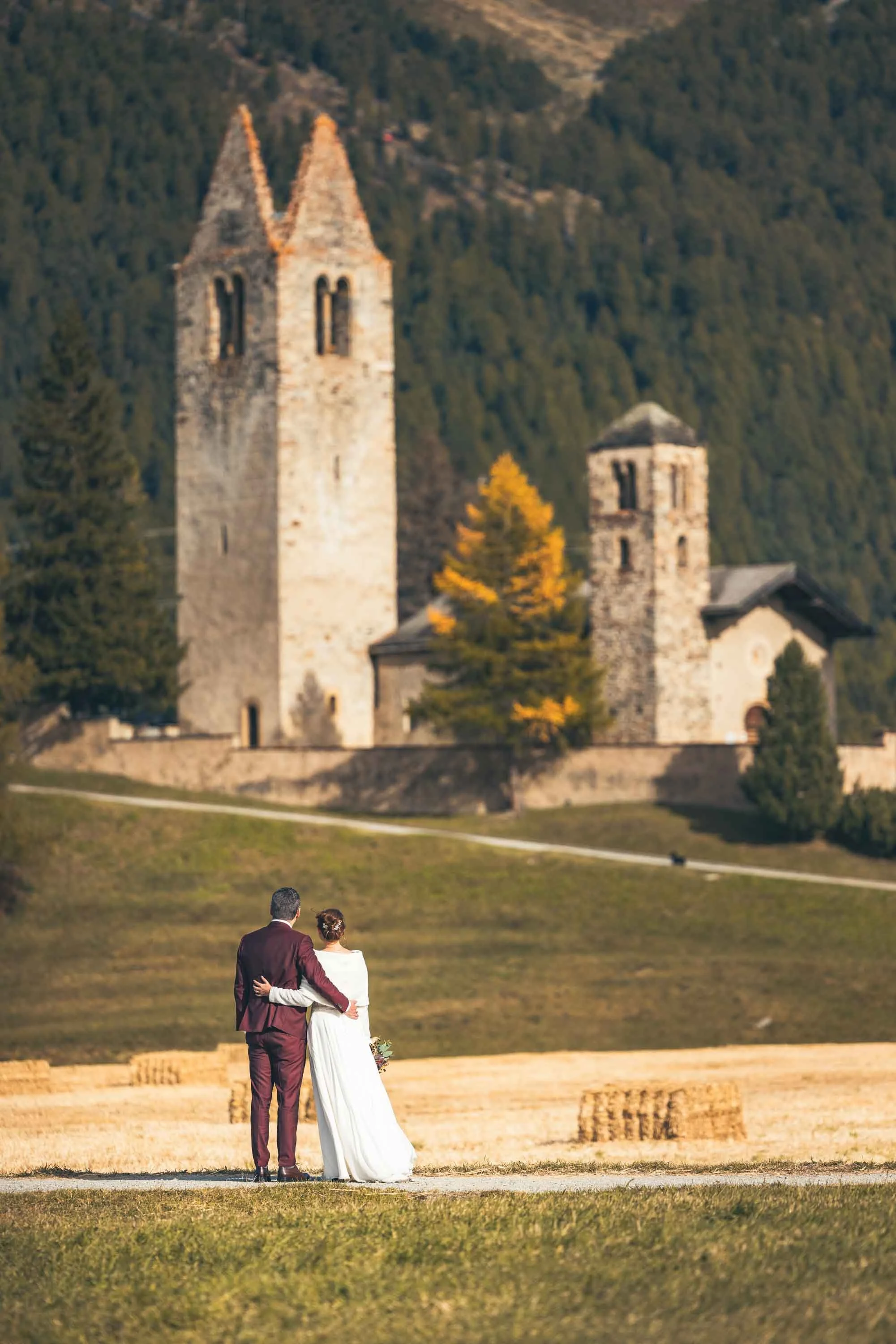 Ein Brautpaar steht auf einer Wiese mit Blick auf eine Burg im Hintergrund, umgeben von Feldern und Bäumen im Herbst.