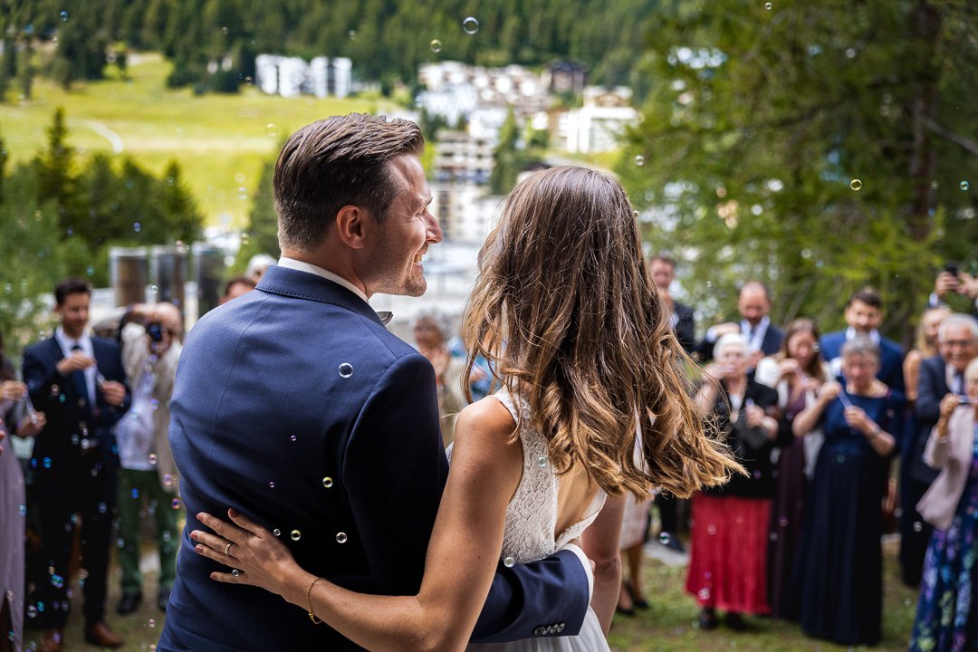 Hochzeitspaar von hinten, im Freien mit Gästen im Hintergrund und Seifenblasen in der Luft.