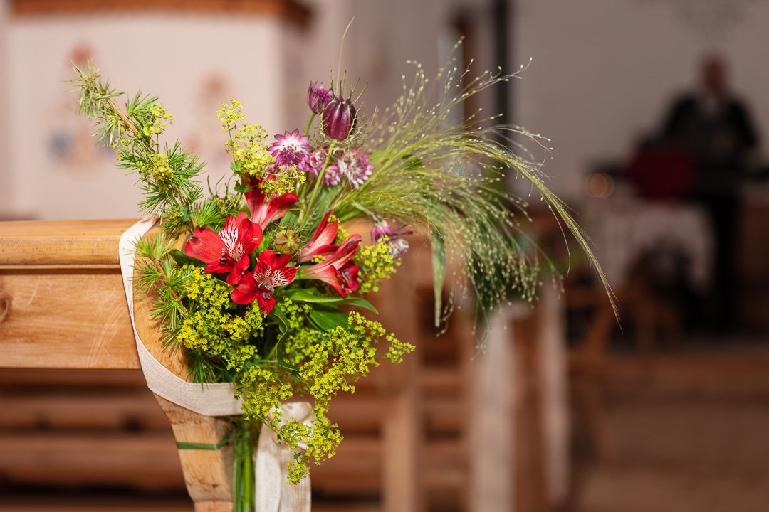 Bunter Blumenstrauß mit roten, gelben und rosa Blumen, befestigt an einer Holzbank in einem Innenraum.