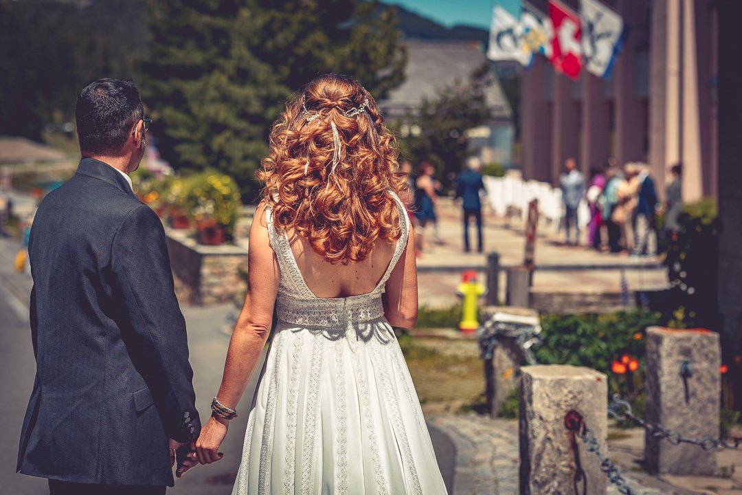 Ein Paar in eleganter Kleidung, die Rückseite eines Mannes im Anzug und einer Frau im Hochzeitskleid, Hand in Hand gehend auf einem Bürgersteig mit Menschen im Hintergrund in feierlicher Atmosphäre.