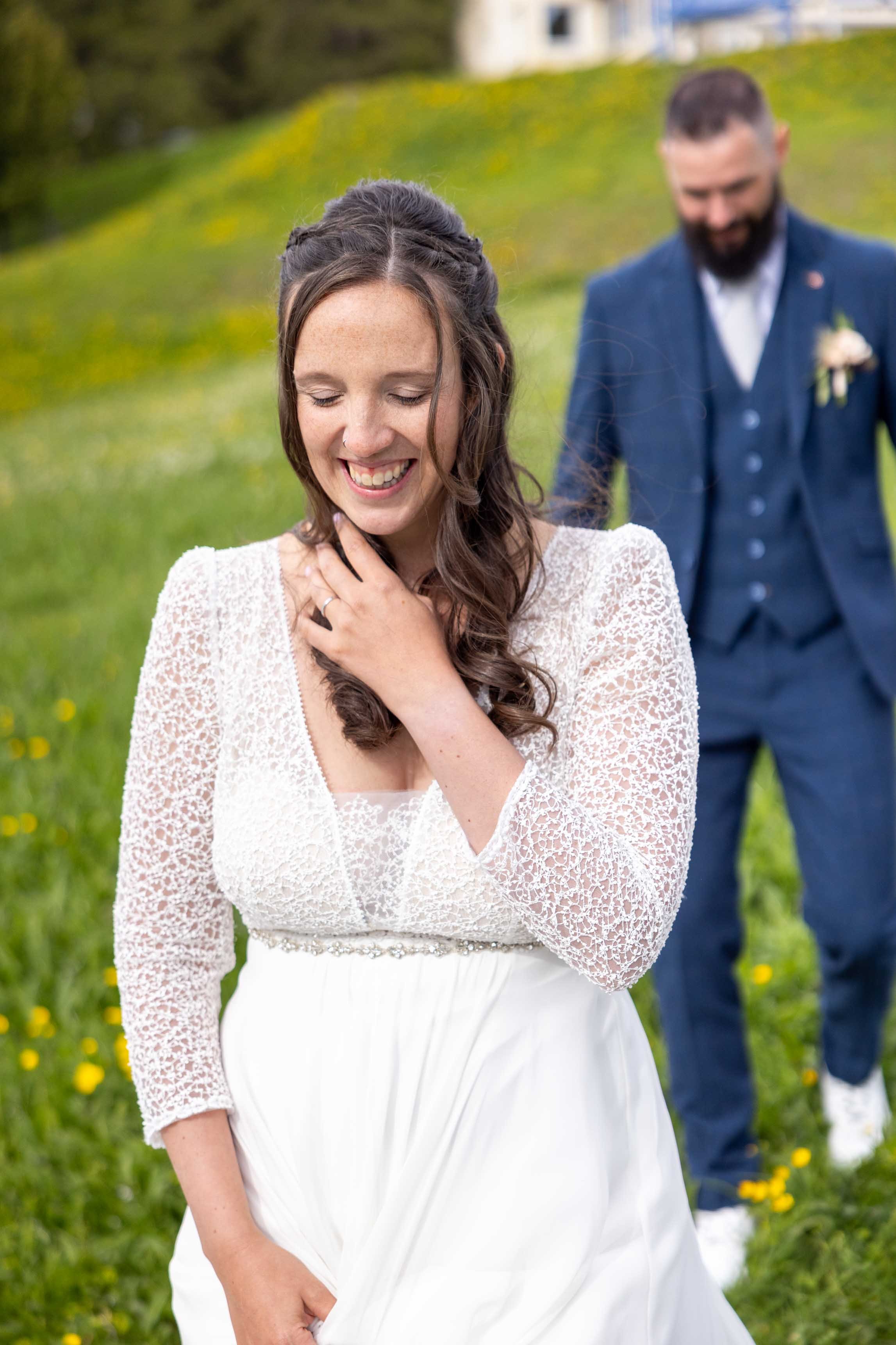 Braut lächelt im Vordergrund, Bräutigam im Hintergrund; Hochzeit im Freien auf einer grünen Wiese.