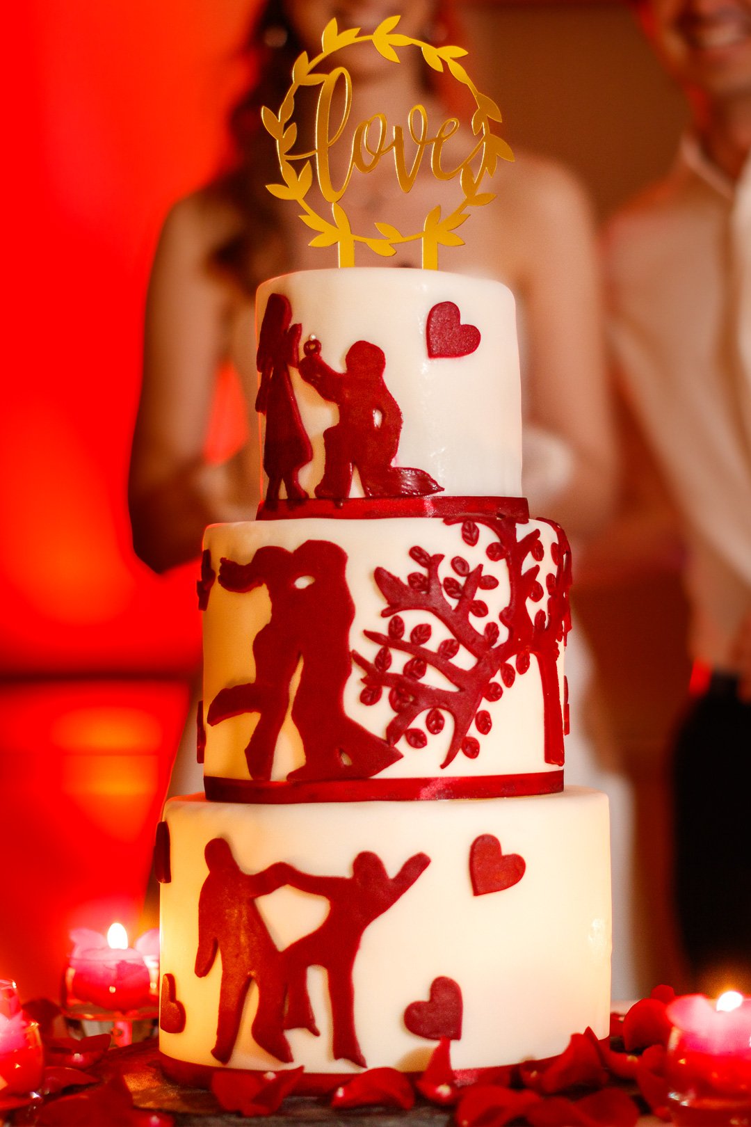 Dreistöckige Hochzeitstorte mit weißen und roten Dekorationen, inklusive Silhouetten von Paaren in roten Herzen und einem "Love"-Topper. Im Hintergrund verschwommene Menschen und rote Beleuchtung.