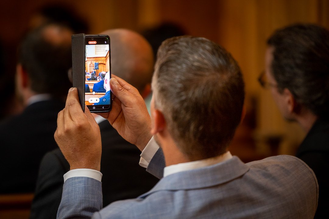 Ein Mann hält ein Smartphone und macht ein Foto oder Video von einem Vortrag oder Event, das auf dem Bildschirm des Handys sichtbar ist. Mehrere Personen sitzen im Hintergrund. Der Mann trägt einen blauen Anzug.