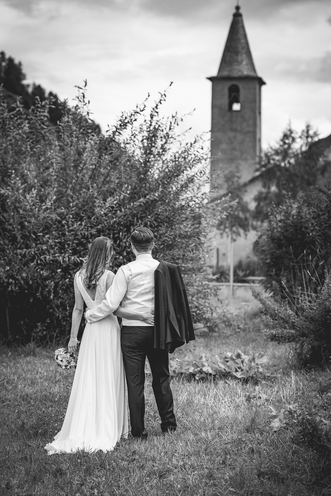 Ein Paar in Hochzeitskleidung steht Rücken an Rücken in einem Garten, eine Kirche im Hintergrund.