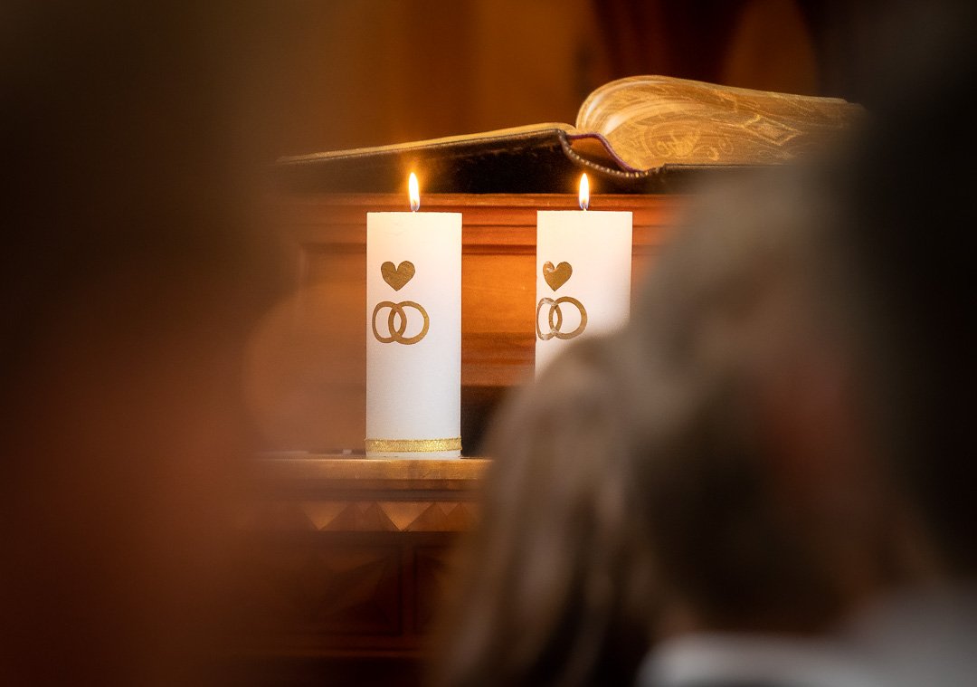 Zwei brennende Hochzeitskerzen mit Herzen und Ringen, daneben eine geöffnete Bibel, unscharfer Vordergrund.