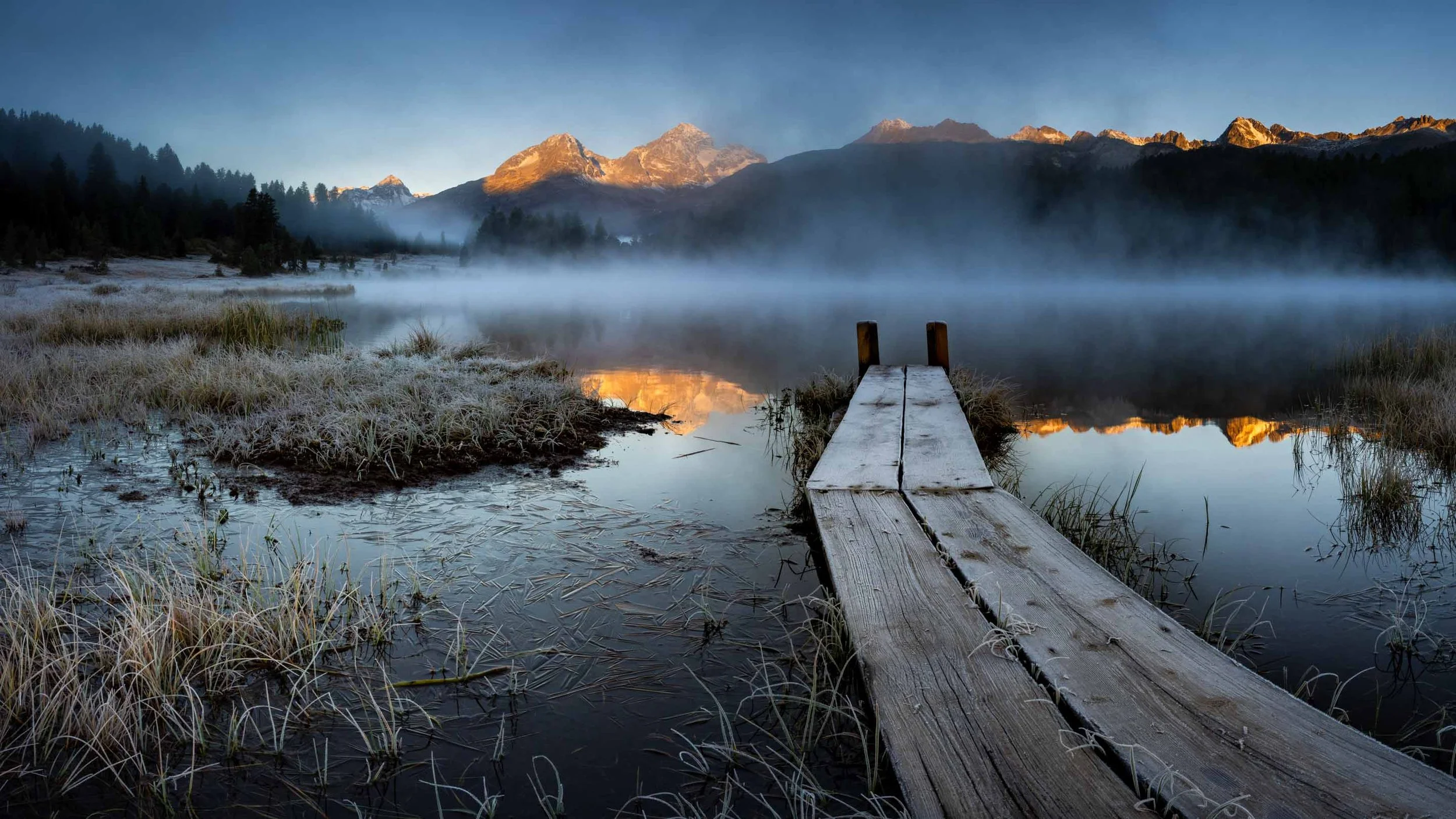 Geschwungener Holzsteg am Stazersee mit Nebel und beleuchteten Gipfeln im Oberengadin.